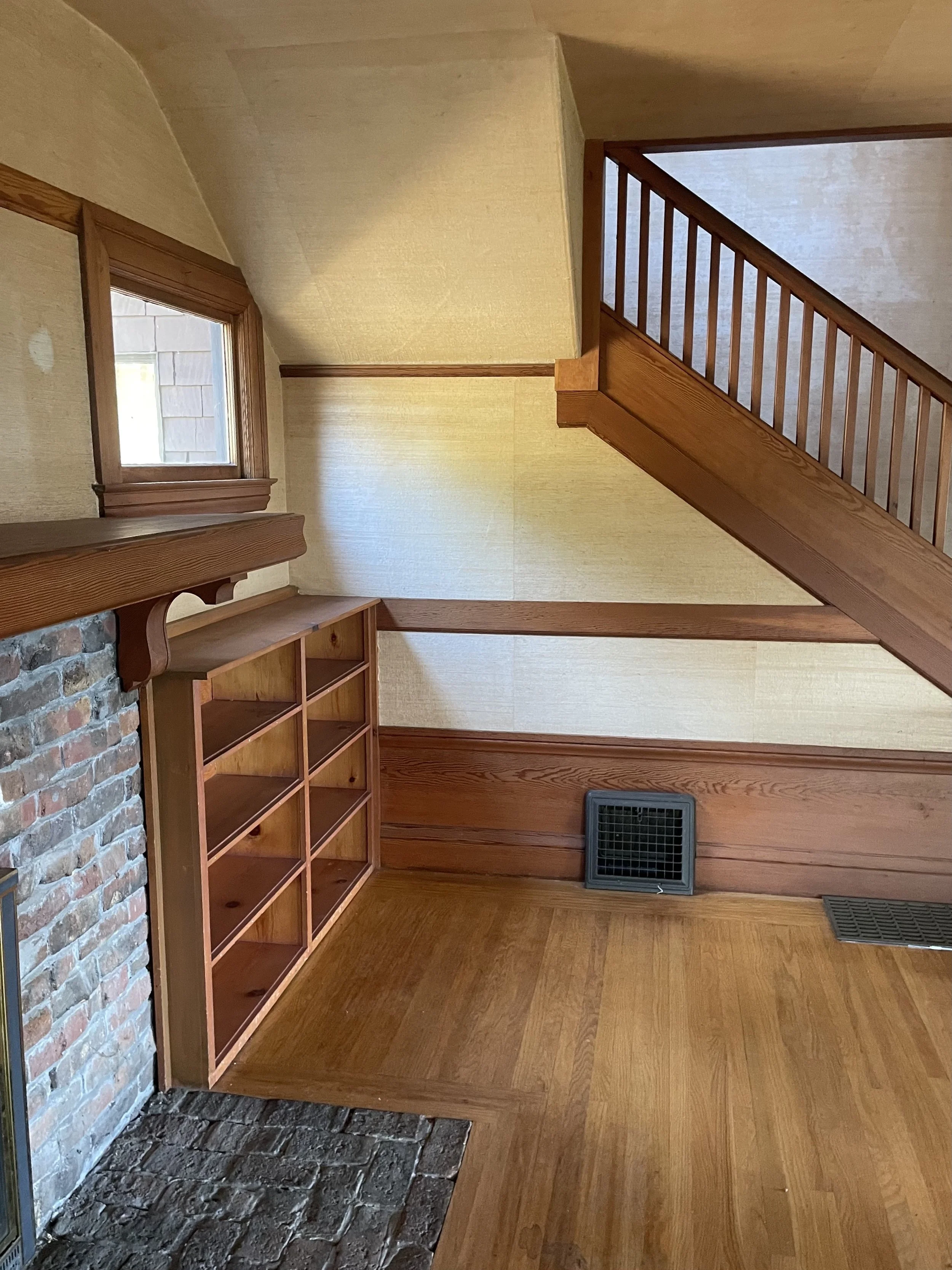 Interior of a room with wood paneling, a brick fireplace, a wooden shelf, and a staircase with a wooden railing.