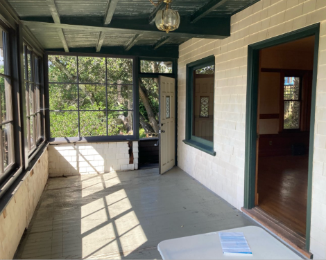 Sunlit enclosed porch with white brick walls, black window frames, and a view of green trees outside. There's a door leading inside and a small table with papers.