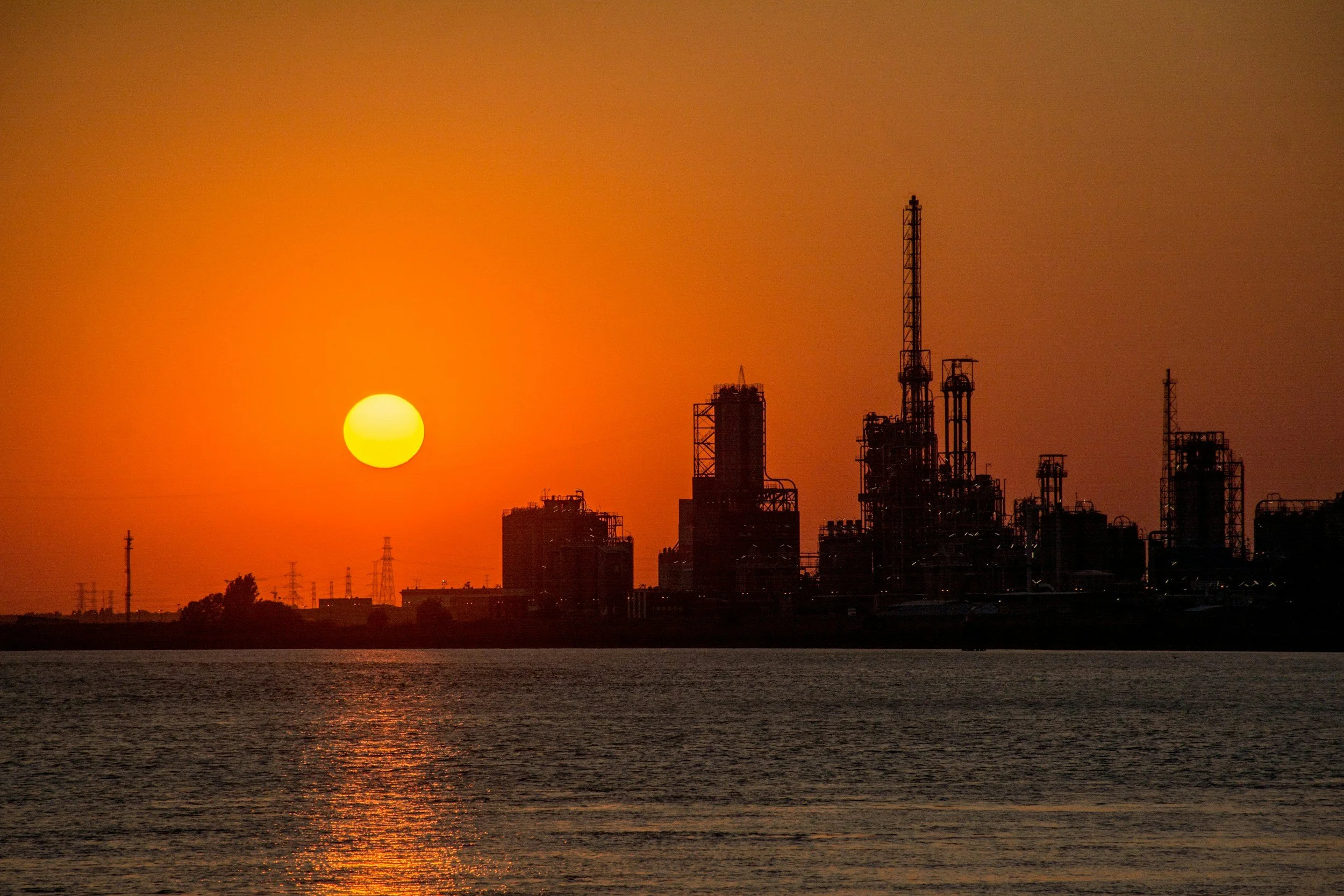 Sunset over an industrial cityscape with silhouettes of factories and communication towers, and a body of water in the foreground.