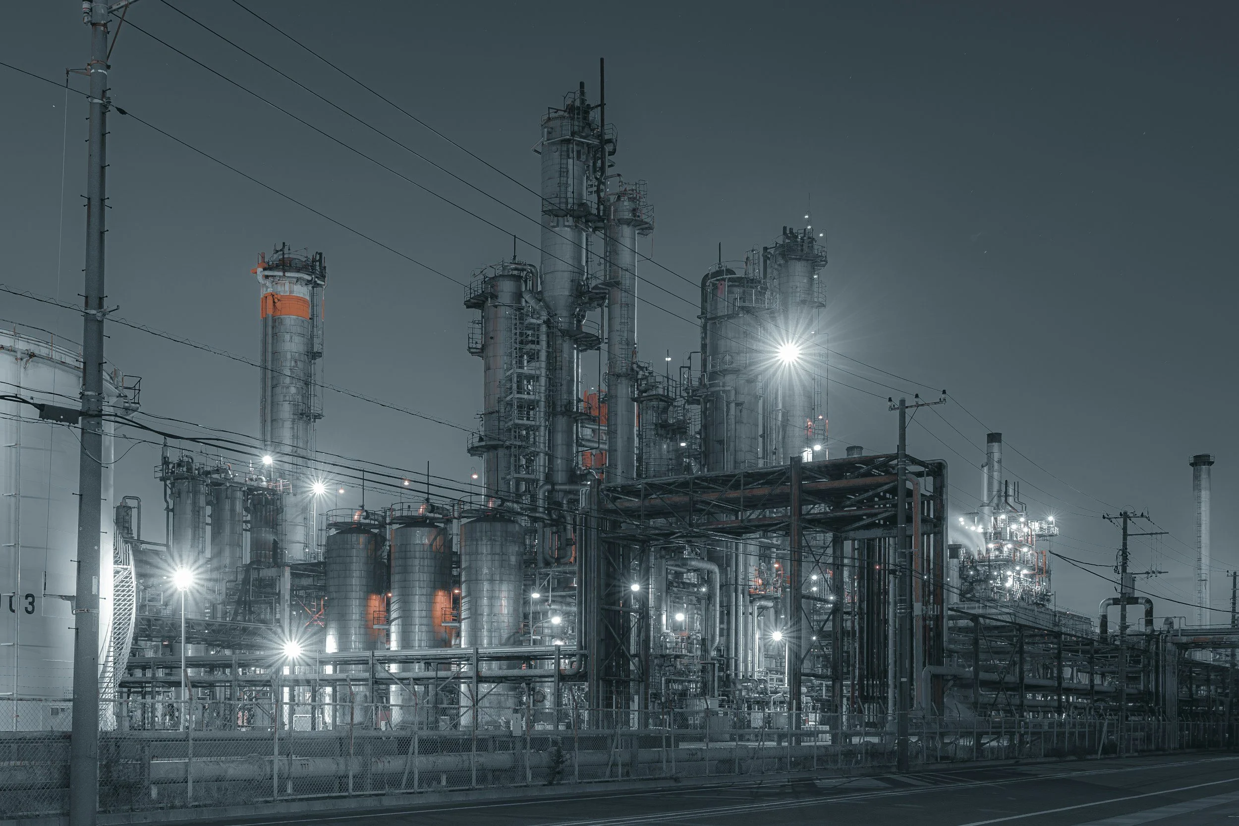 Industrial refinery with multiple tall metallic structures, pipes, and electrical wires illuminated by bright lights at night.