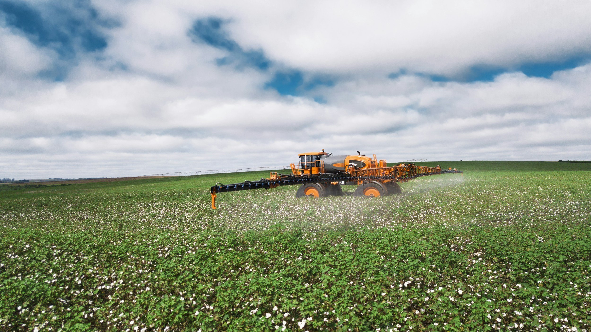 A large orange tractor spraying crops in a green field under a cloudy sky.