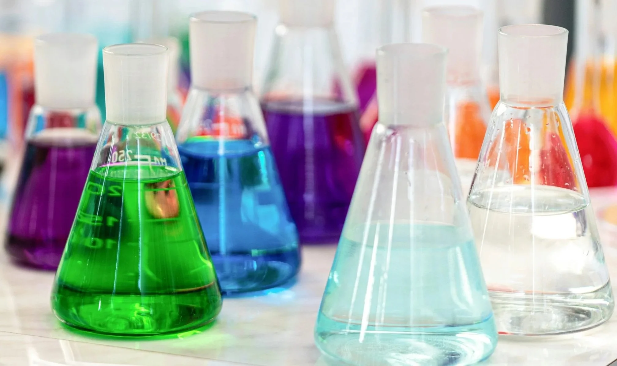 Several laboratory flasks with colorful liquids including green, blue, purple, and clear, arranged on a lab bench.