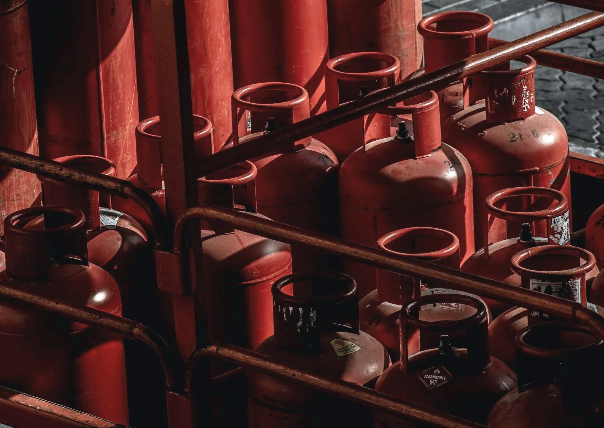 A close-up of many red propane gas cylinders stored outdoors on a rack, with some labels and tags visible on them.