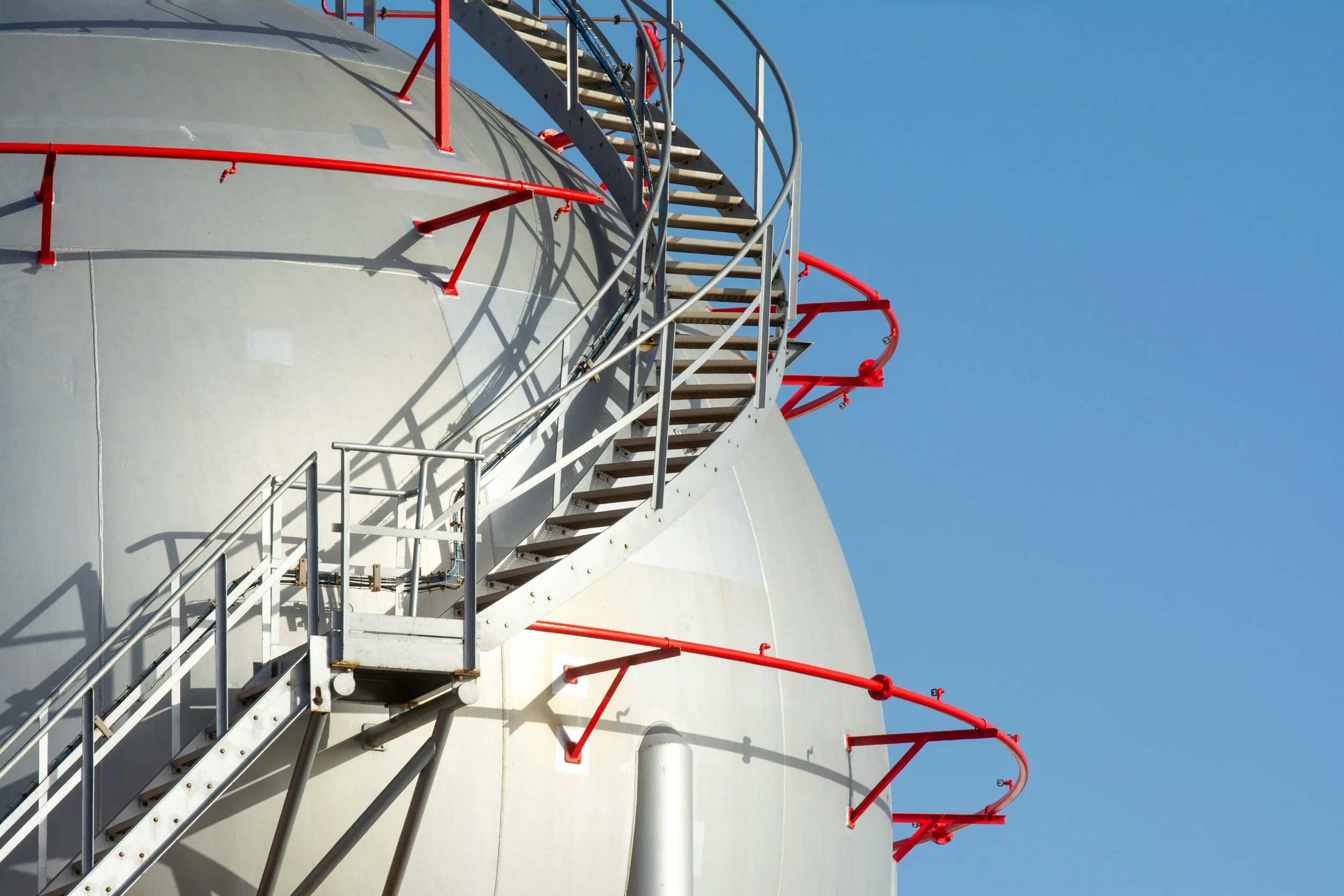 A white industrial storage tank (sphere) with red safety railings and a spiral staircase, against a clear blue sky.