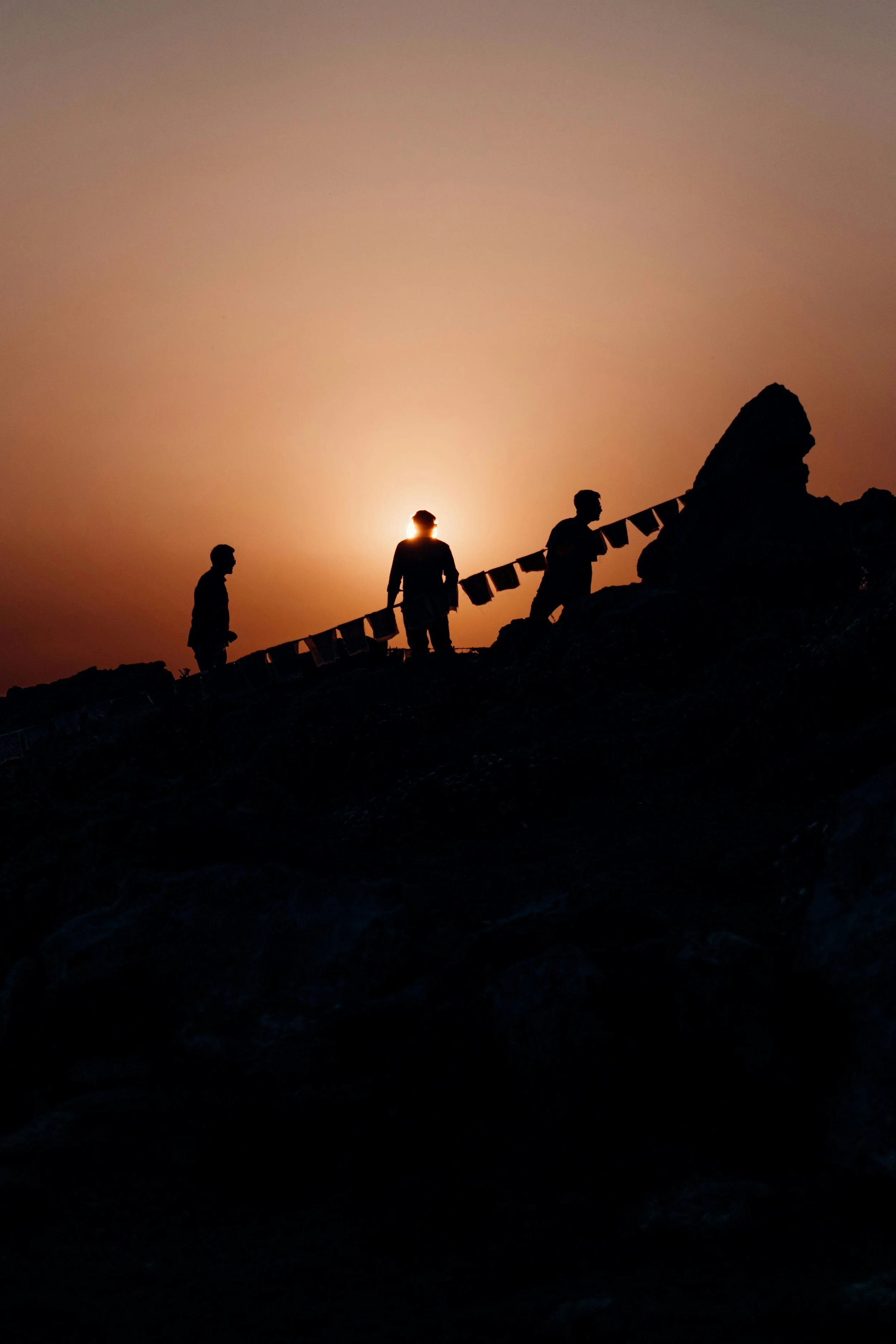 Silhouettes of three people walking on a rocky terrain at sunset, with one person holding a string of prayer flags, and a large rock formation on the right side of the image.