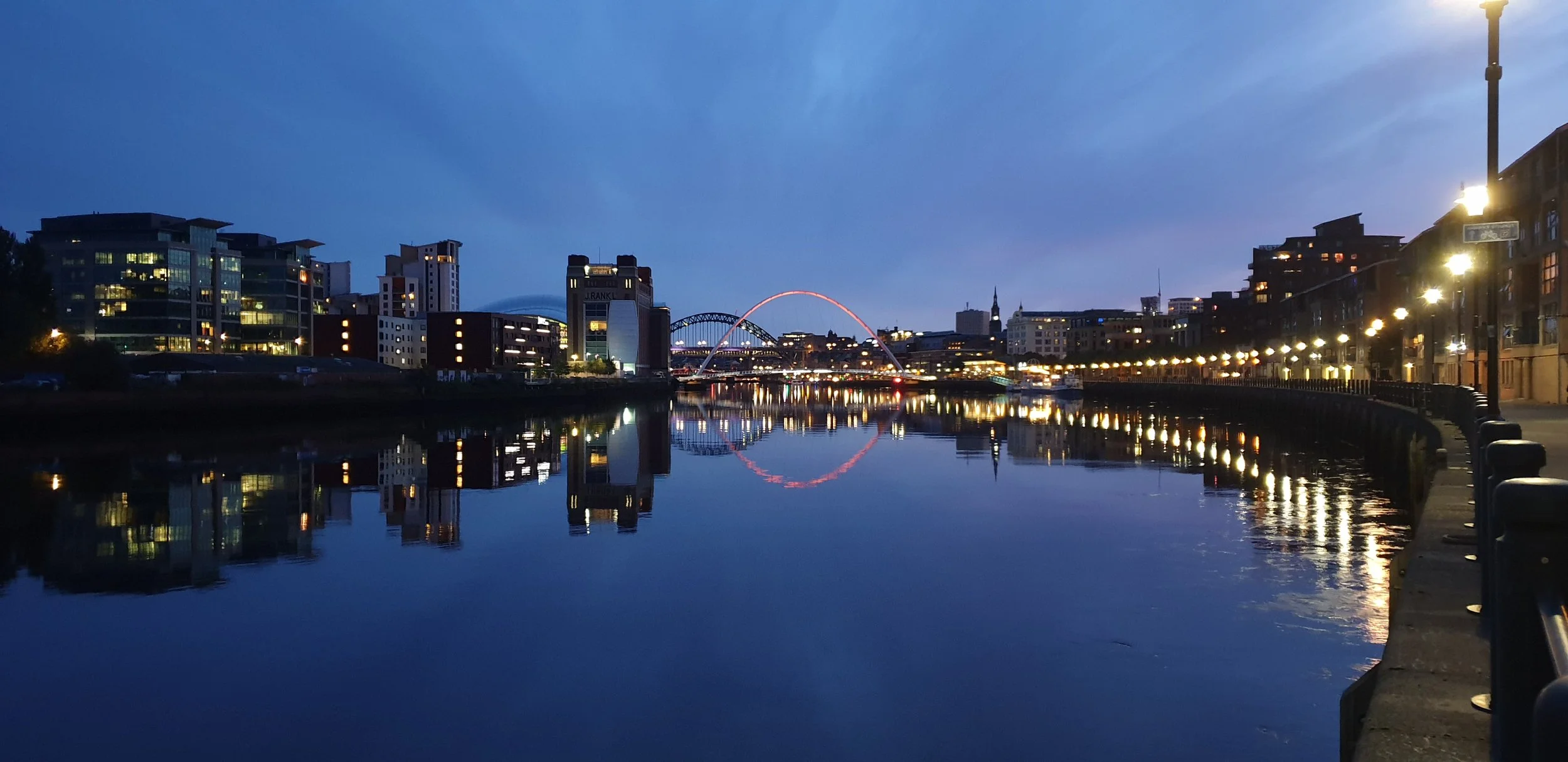 Cityscape at dusk showing buildings, a bridge with lights, and a ferris wheel reflected in calm water along a waterfront promenade.