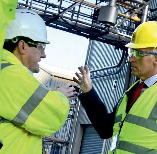 Two male construction workers wearing yellow hard hats and high-visibility safety vests having a discussion at an industrial site.