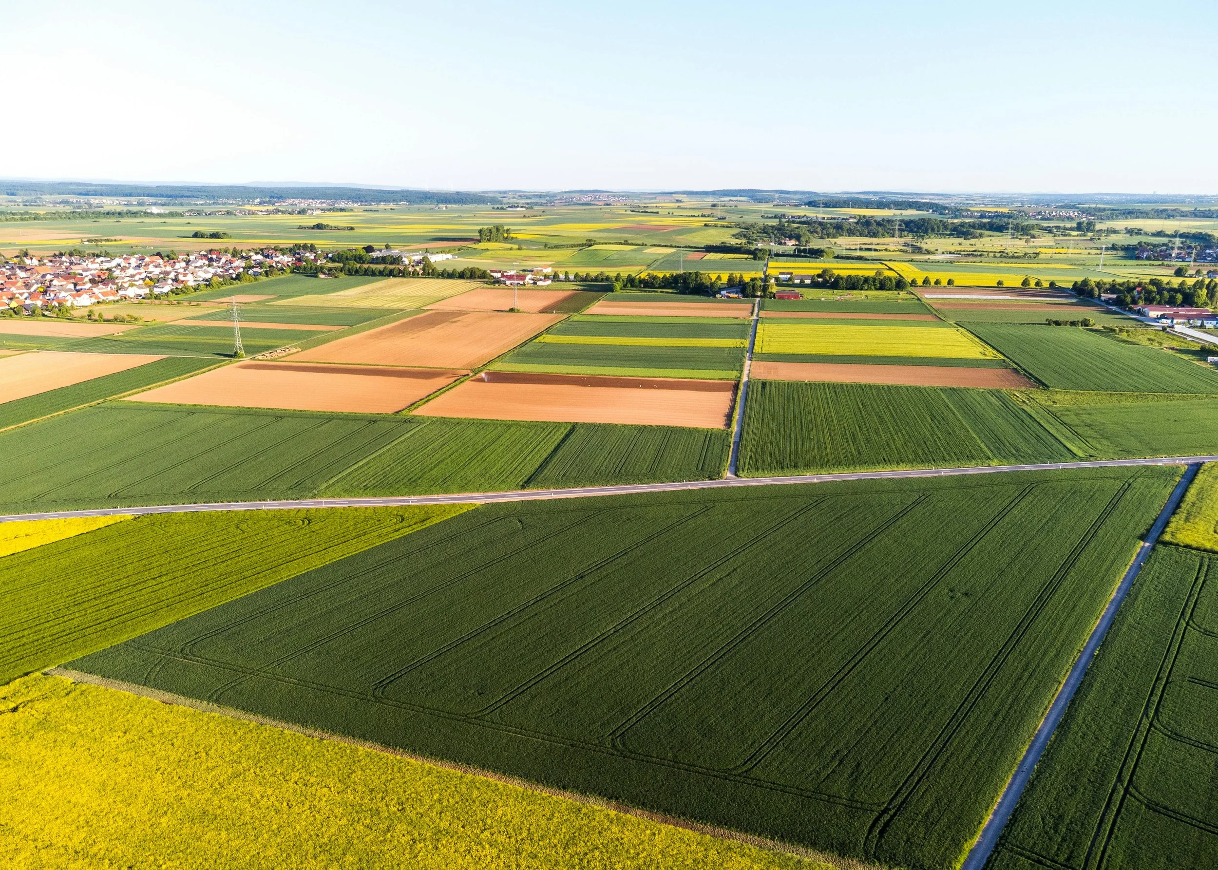 Aerial view of a rural landscape with colorful agricultural fields, a small village with houses, and patches of green trees under a clear sky.