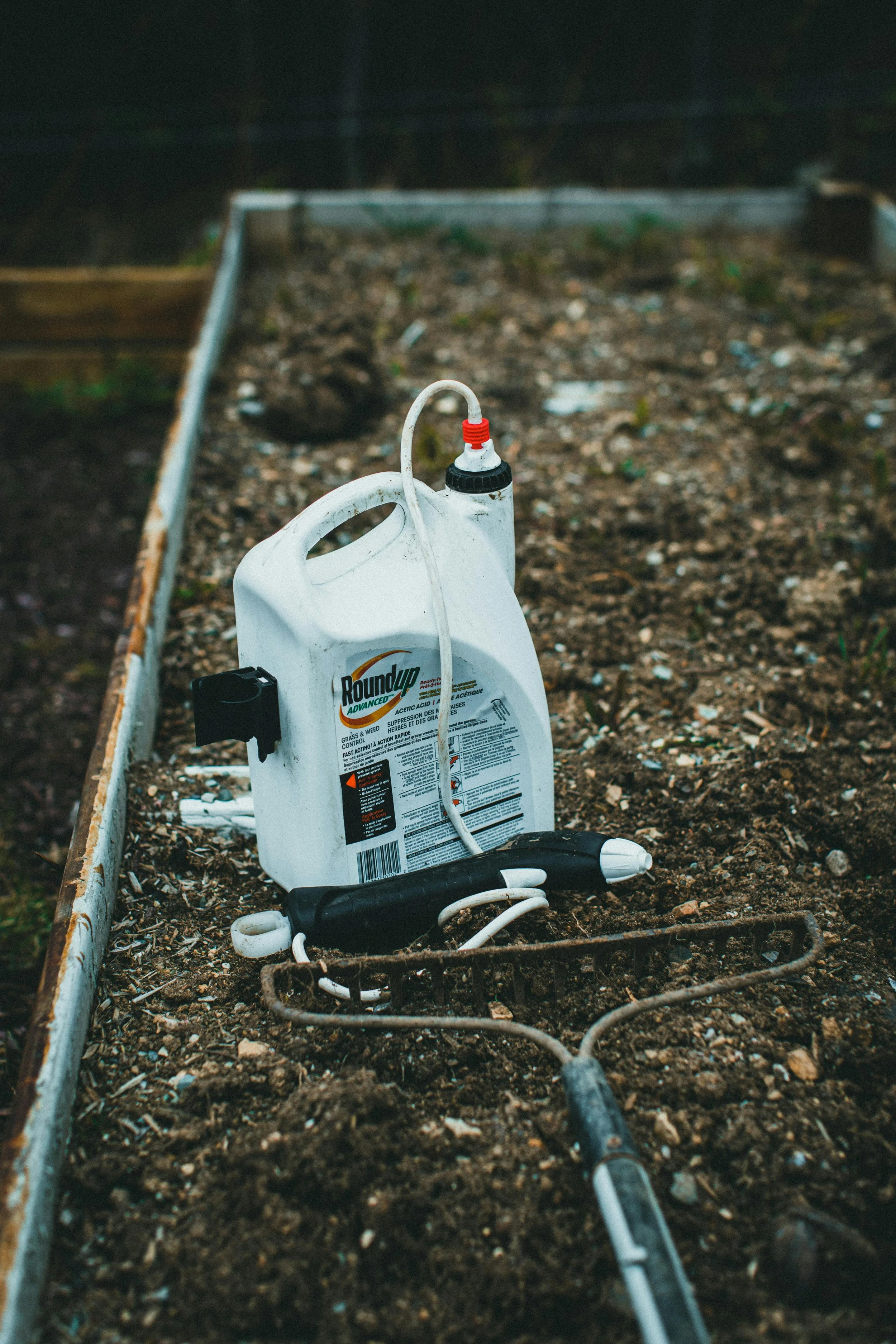 A garden bed with a bottle of Roundup weed killer and a hose, with dirt and small rocks, surrounded by a metal border.