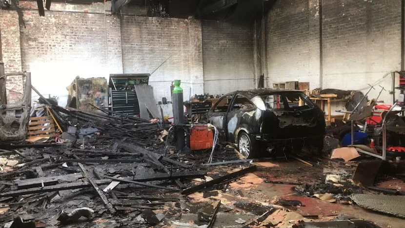 Interior of a building damaged by fire, with a burnt-out car in the center, charred debris scattered on the floor, and broken furniture and equipment around.