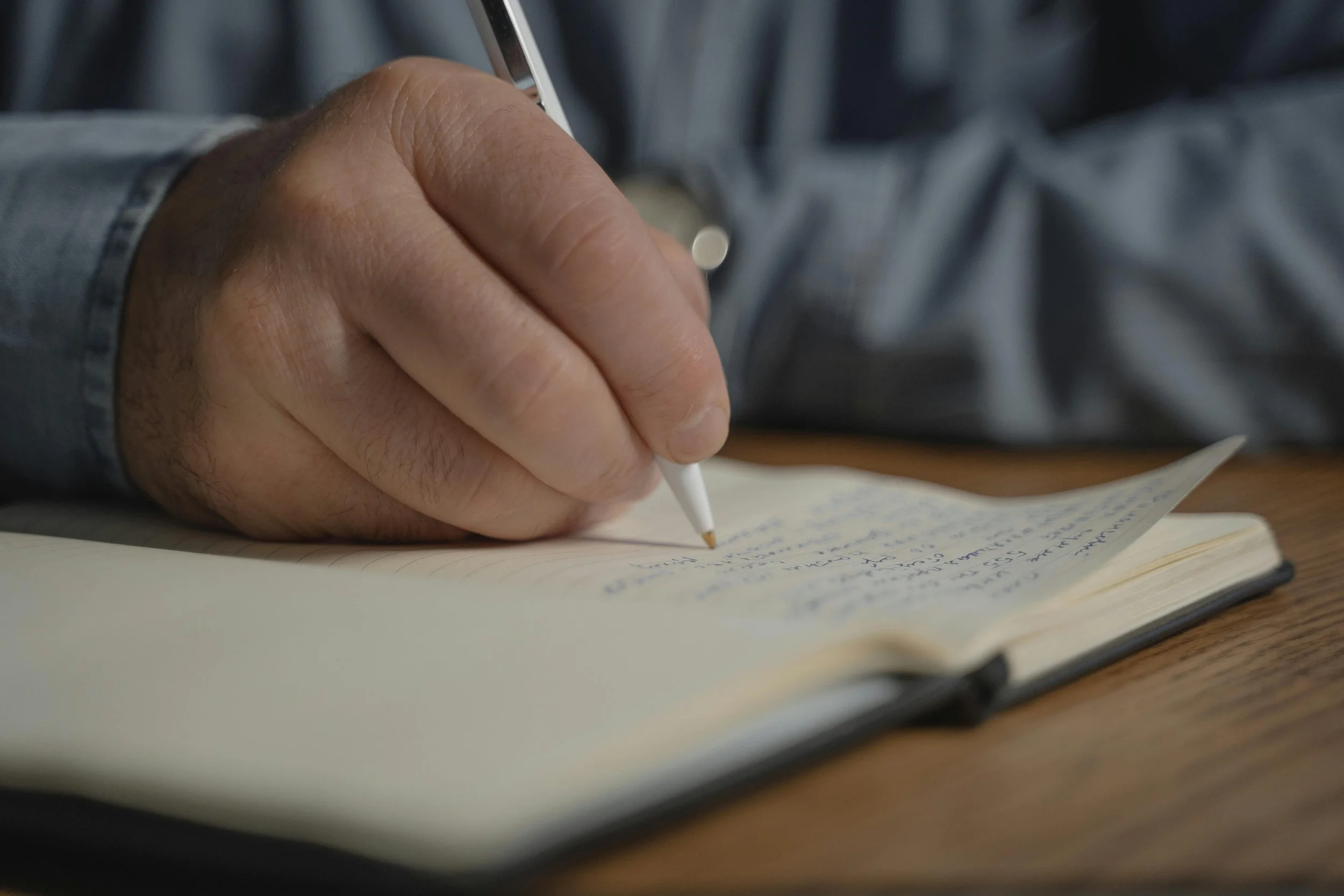 Close-up of a hand holding a white pen and writing in a notebook with handwritten notes, on a wooden surface.