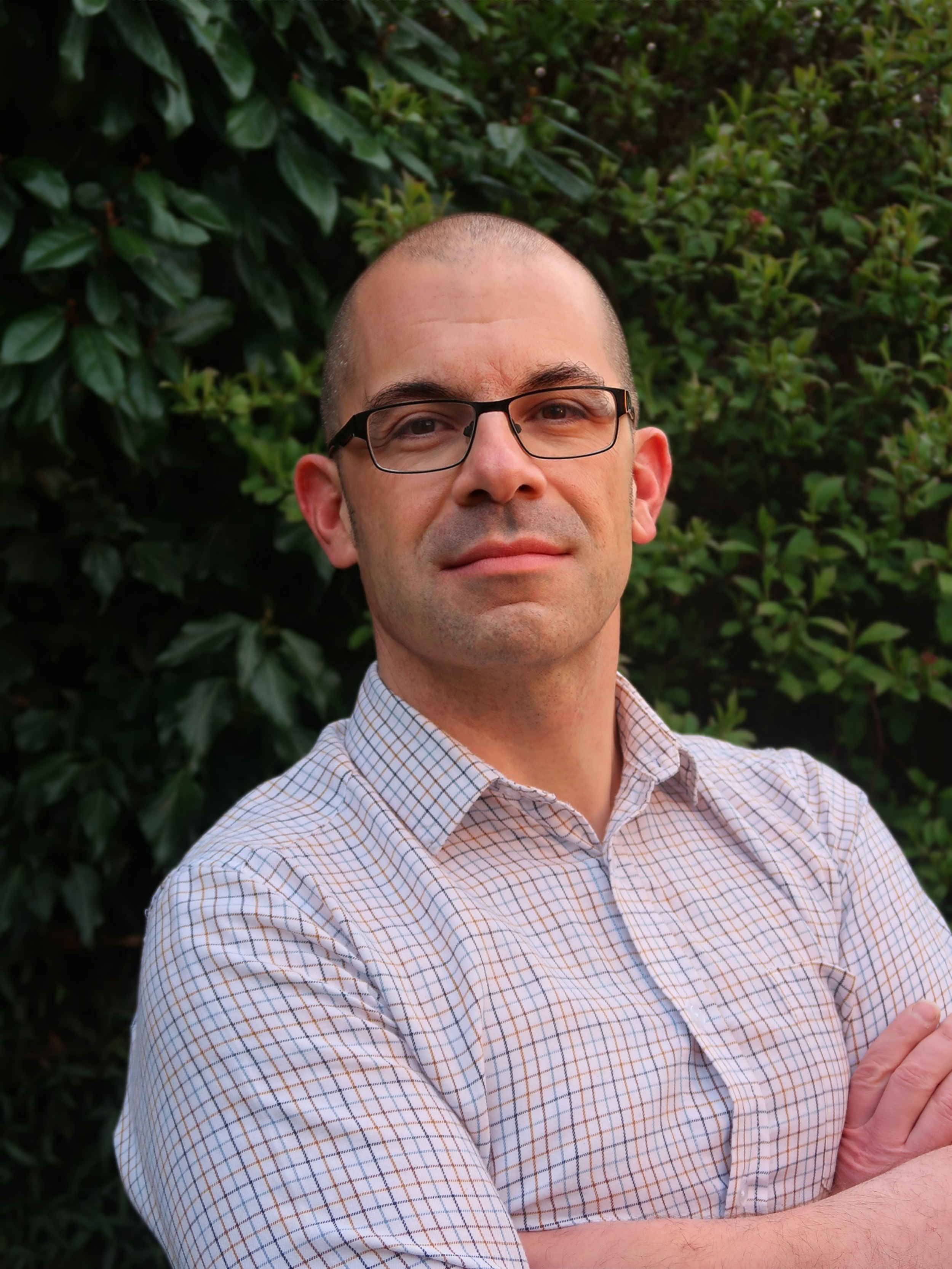 A man with glasses and a checkered shirt standing outdoors in front of green foliage, crossing his arms.