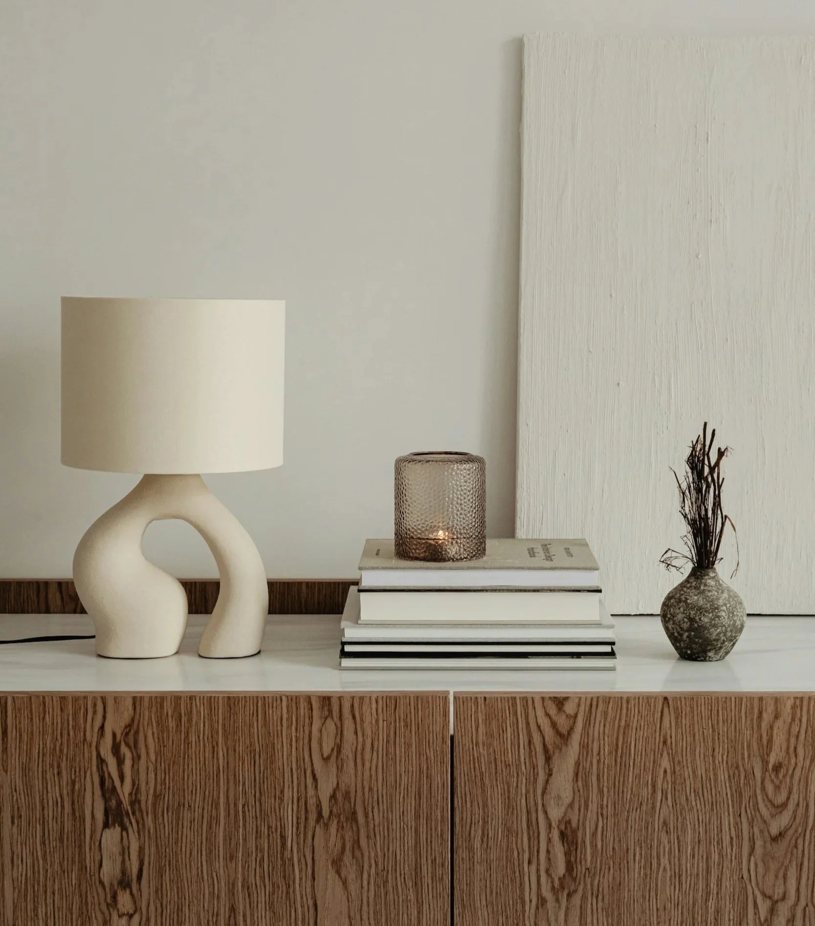 Decorative sideboard with a modern beige lamp, a stack of books, a candle in a textured glass holder, and a small vase with dried twigs.