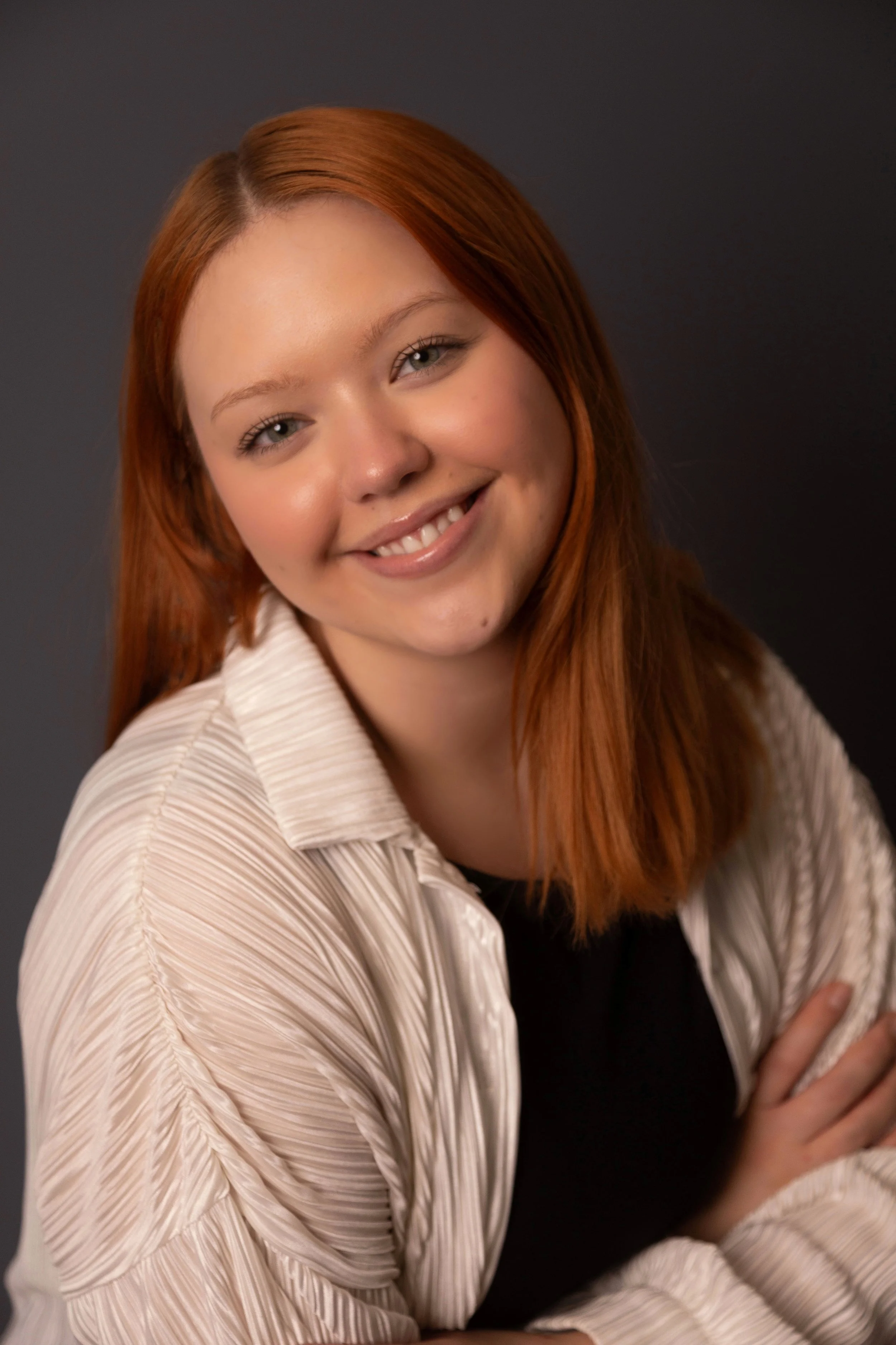 A young woman with red hair smiling and wearing a beige textured jacket over a black top, posed against a dark gray background.