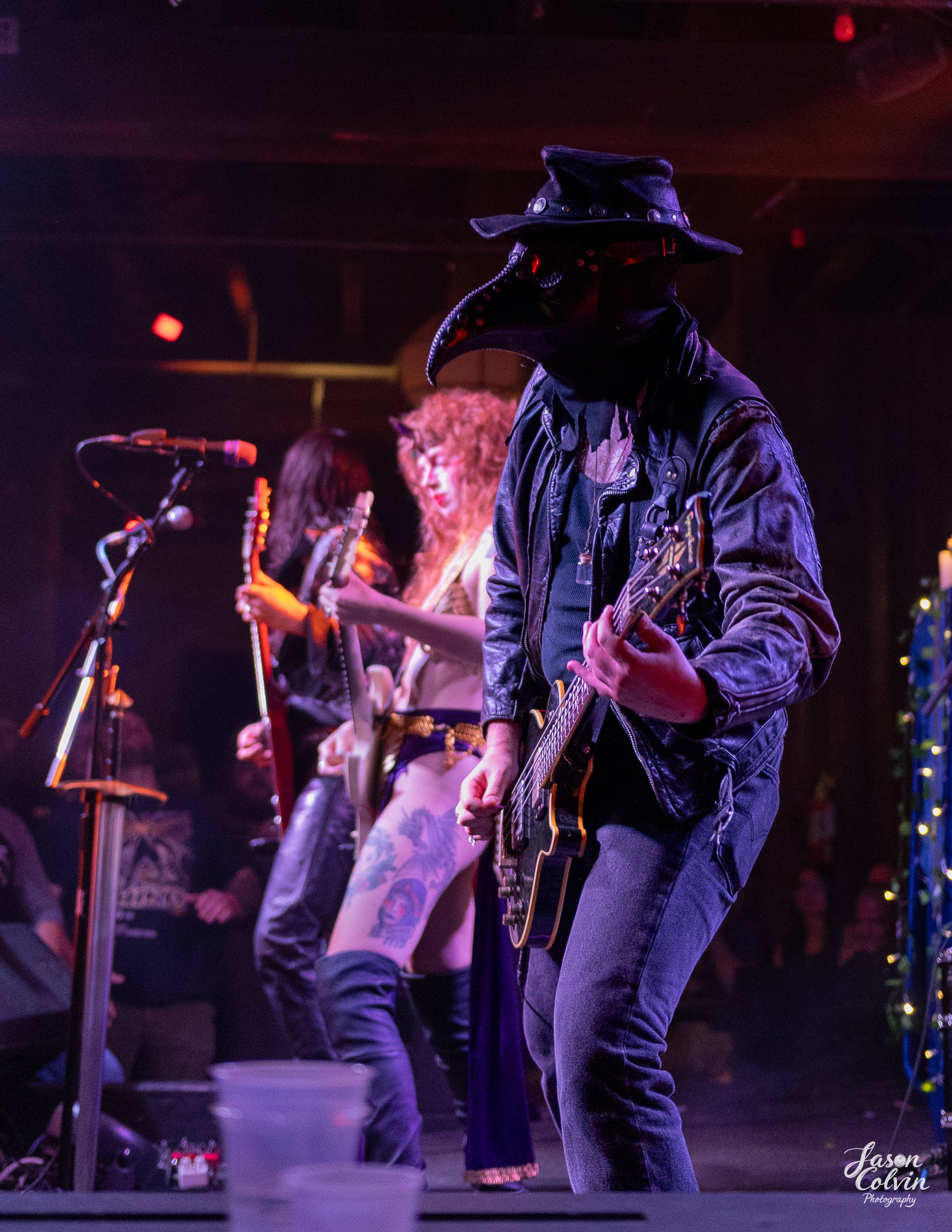 Musicians performing on stage, including a guitarist wearing a plague doctor mask and hat, a woman with curly hair and tattoos, and another woman holding a tambourine, with colorful stage lighting.