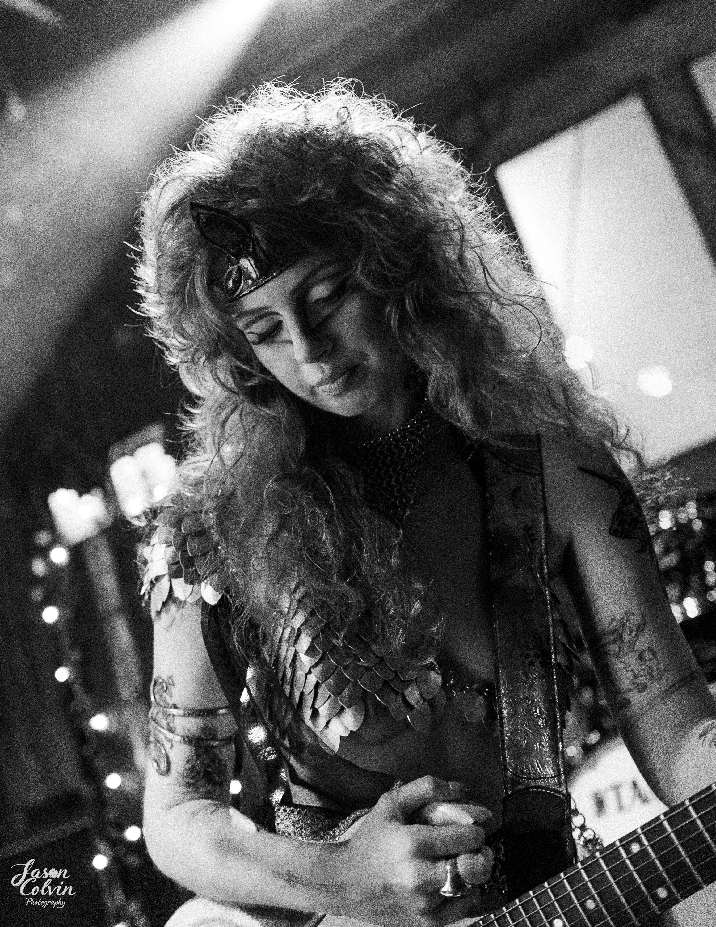 Black and white photo of a woman with curly hair wearing a cat-shaped headband, metallic scale-like clothing on her shoulders, and tattoos on her arm, holding a guitar in a dimly lit setting.