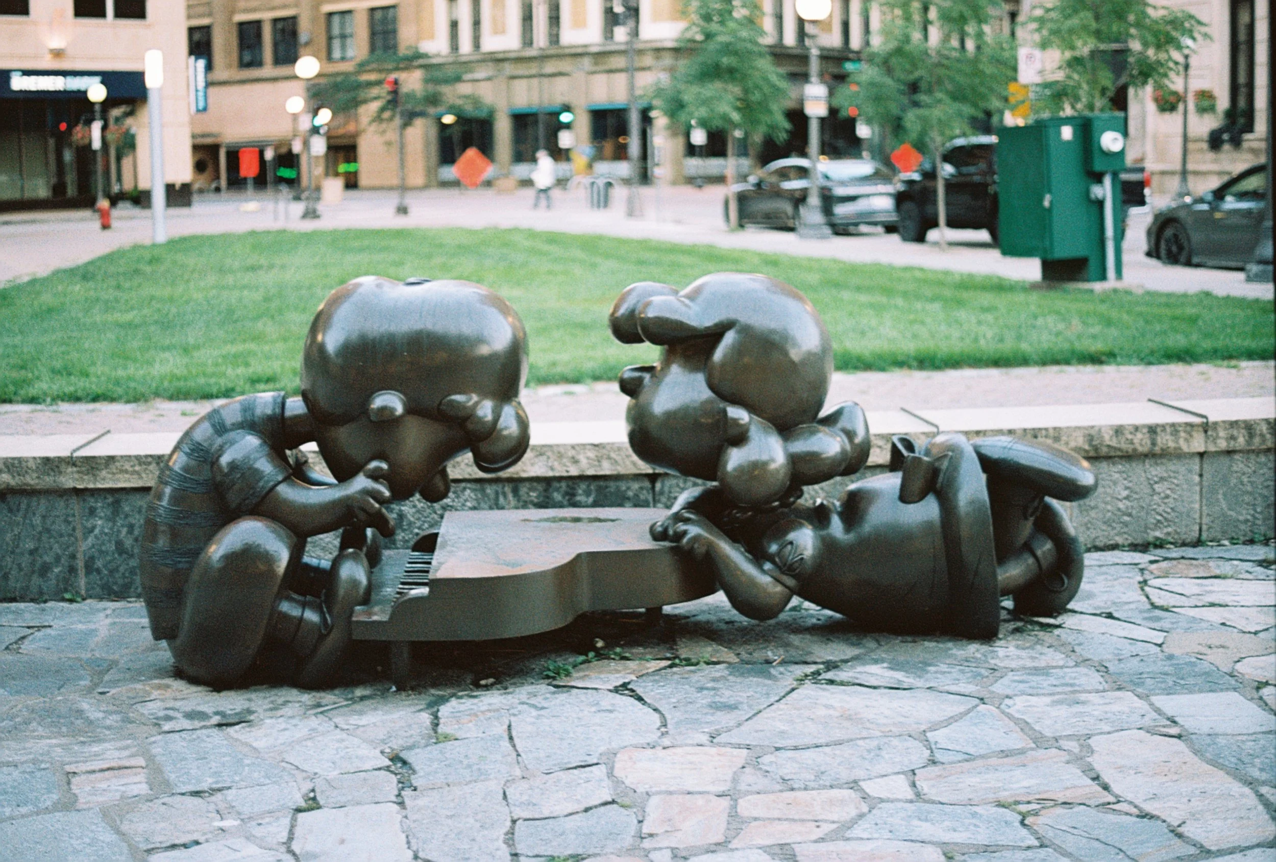 Bronze sculpture of two children, one is playing a piano, on a sidewalk in an urban park.