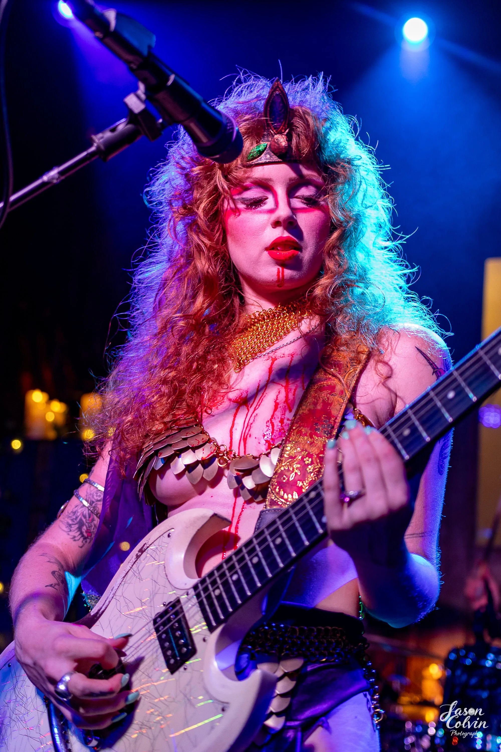 A woman with curly, colorful hair playing an electric guitar on stage, wearing a crown and decorative jewelry, with dramatic makeup and blood-like streaks on her face and chest.