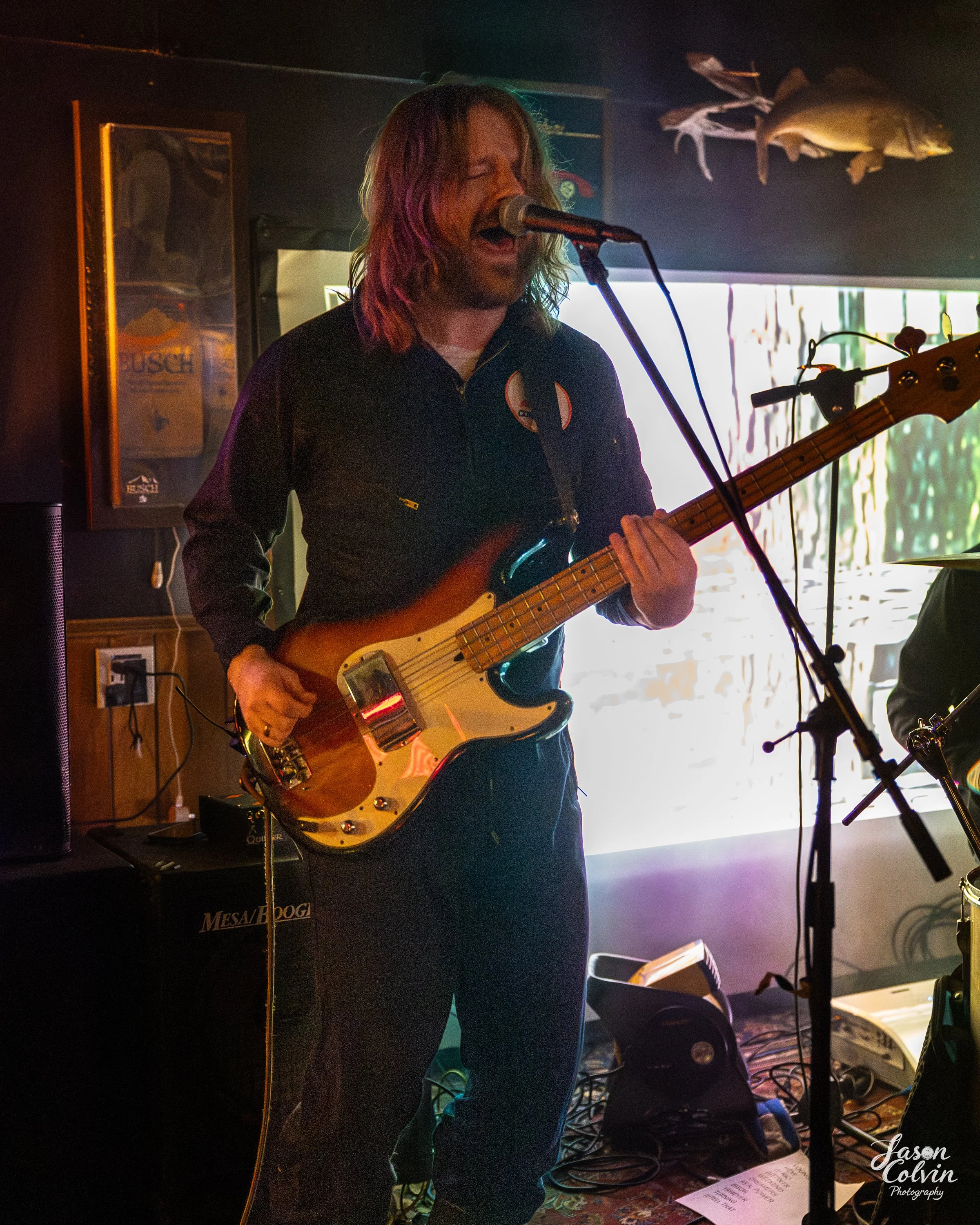 A man with long hair wearing a black jacket is playing electric guitar and singing into a microphone in a dimly lit room. Fish mounted on the wall and a TV screen are visible in the background.