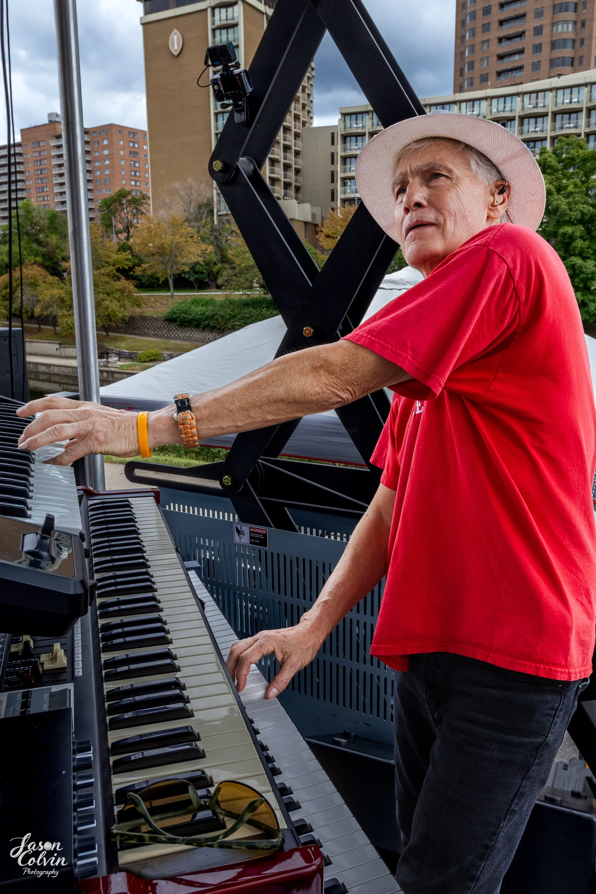 Older man with gray hair, wearing a beige hat and a red shirt, playing a keyboard outdoors with city buildings and trees in the background.