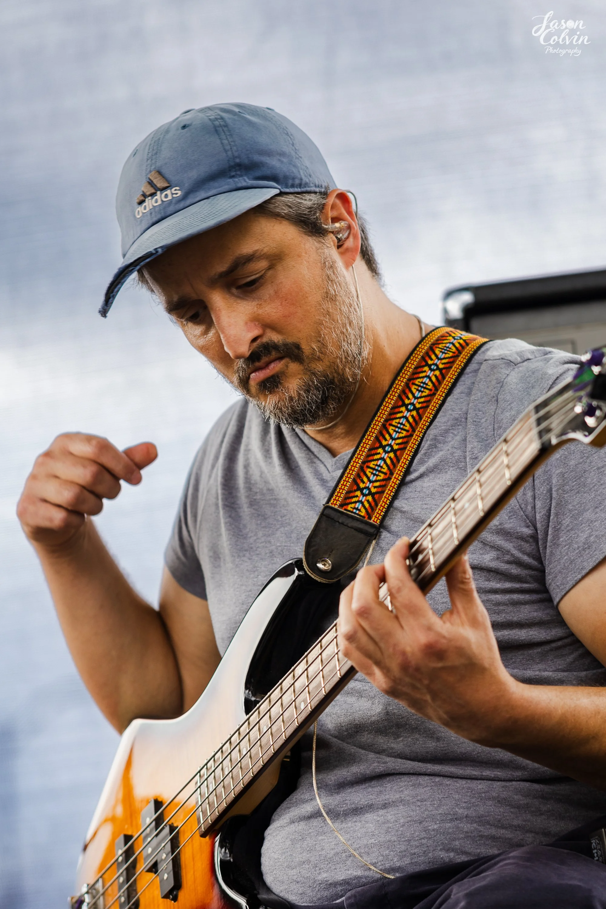 Man playing an electric bass guitar, wearing a blue cap and gray t-shirt, with a colorful strap, in a music practice space.