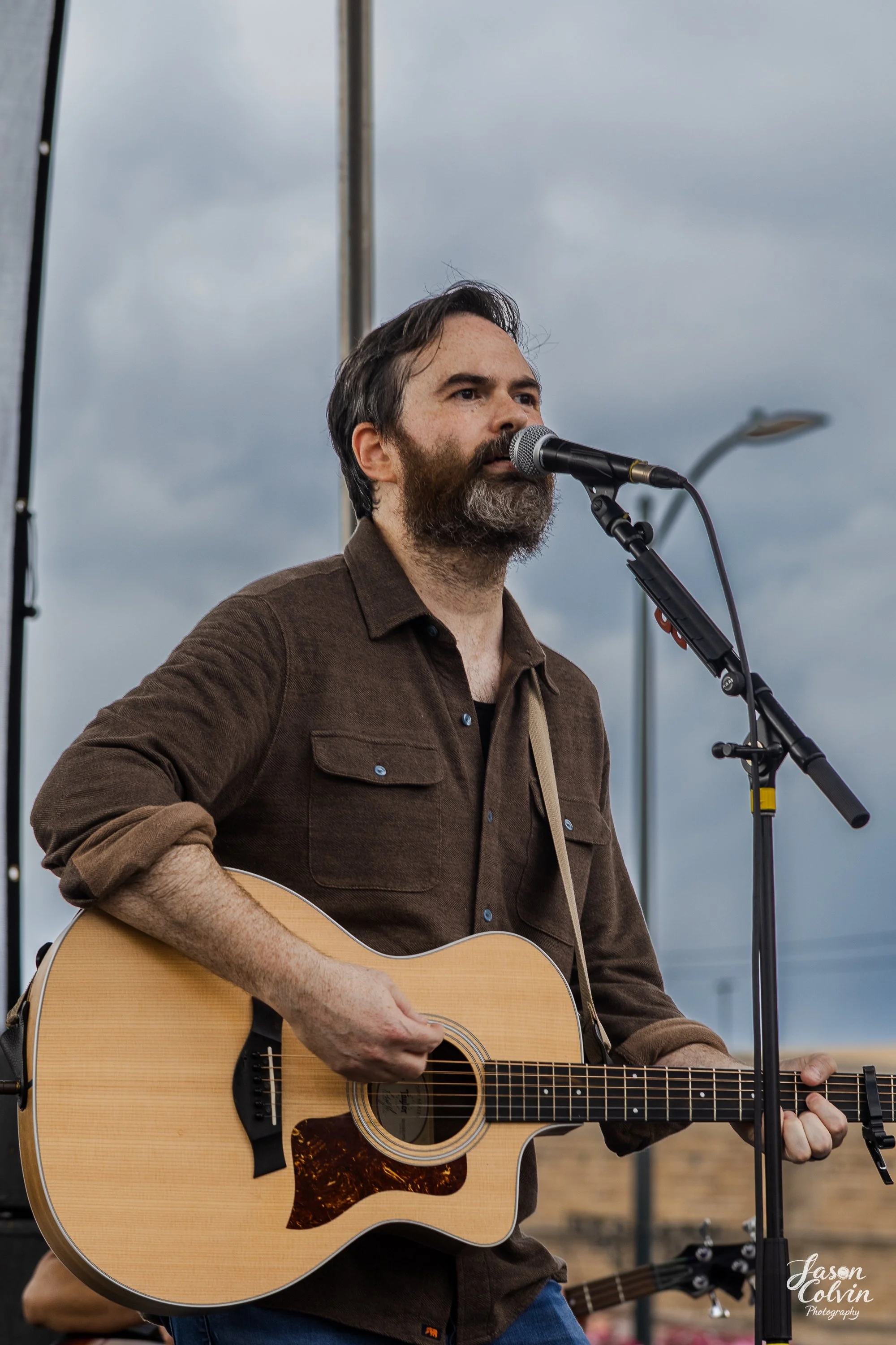 A man with a beard playing an acoustic guitar and singing into a microphone on an outdoor stage on a cloudy day.