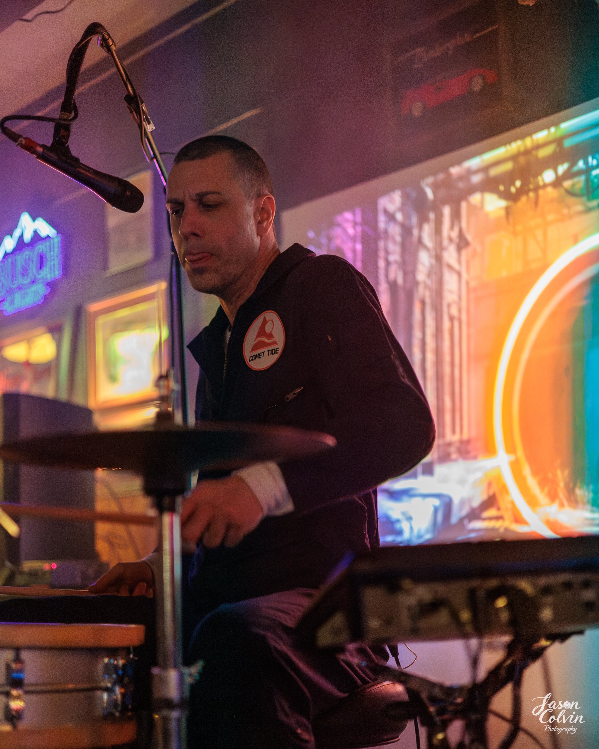 A person playing drums in a dimly lit room with neon signs and colorful lighting in the background.