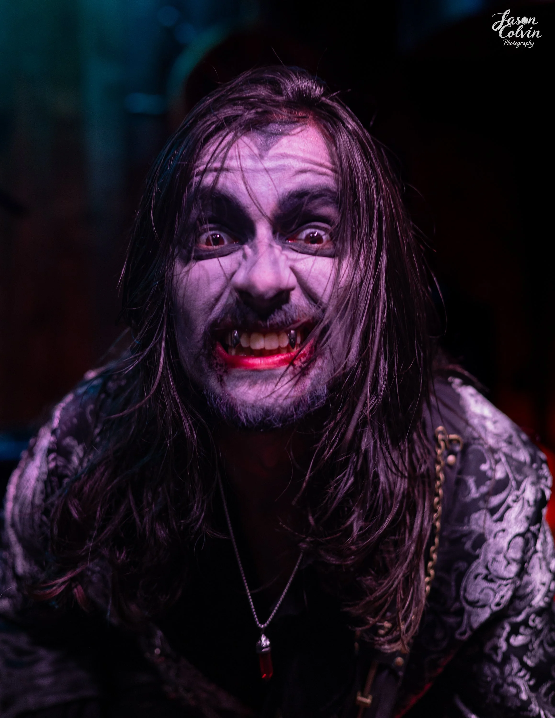 Man with long hair, vampire makeup, fangs, and a necklace, smiling in a dark setting with purple and red lighting.