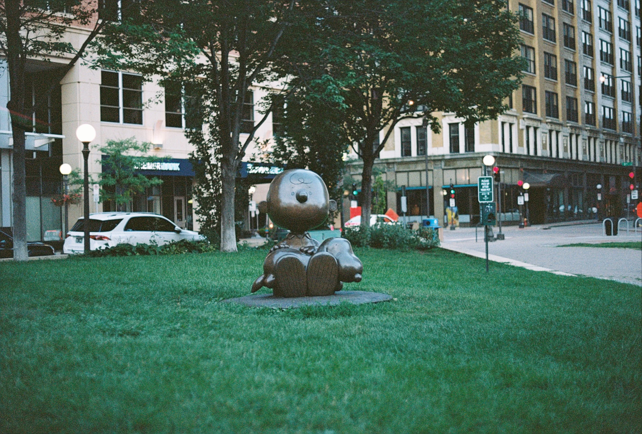A park with a bronze statue of a child sitting on a dog, surrounded by green grass, trees, and city buildings in the background.