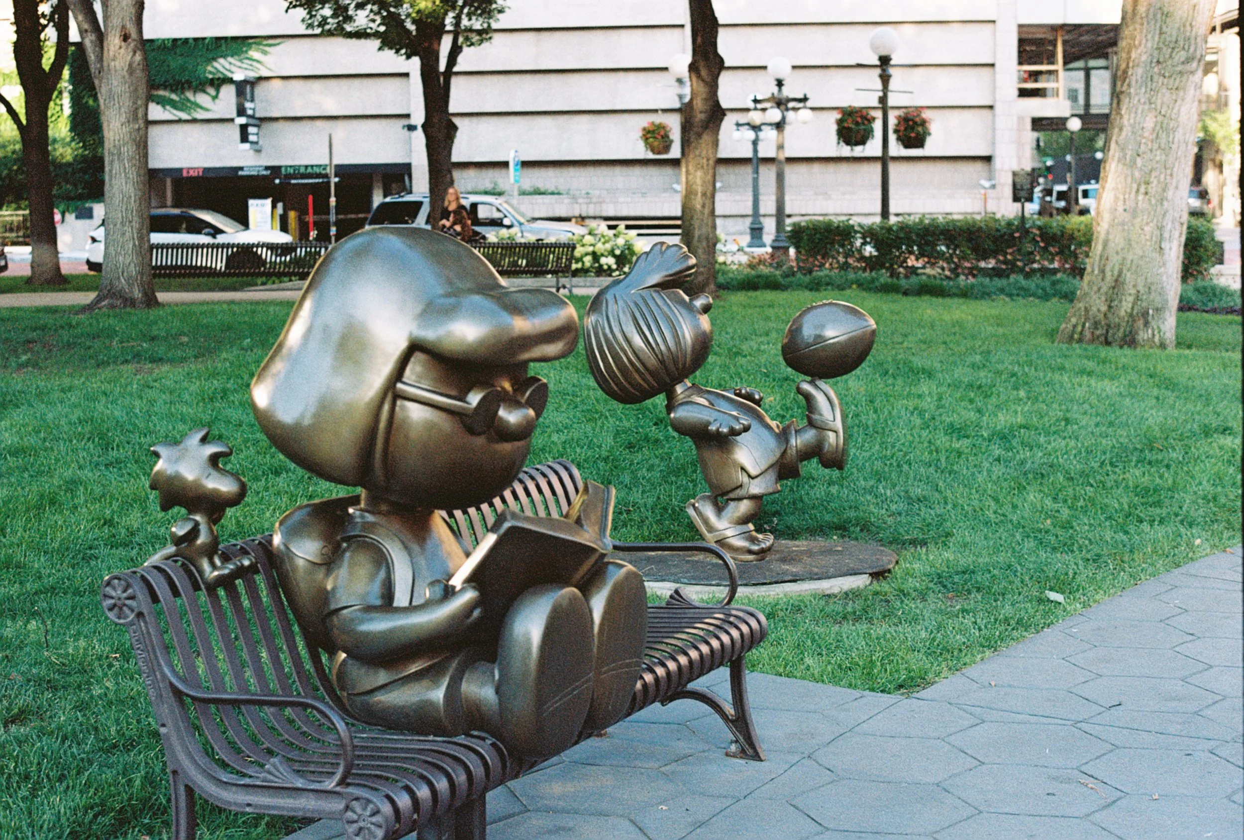 Bronze statues of two children, one sitting reading a book on a park bench and the other walking with a ball in hand, in a green park with trees and a buildings in the background.