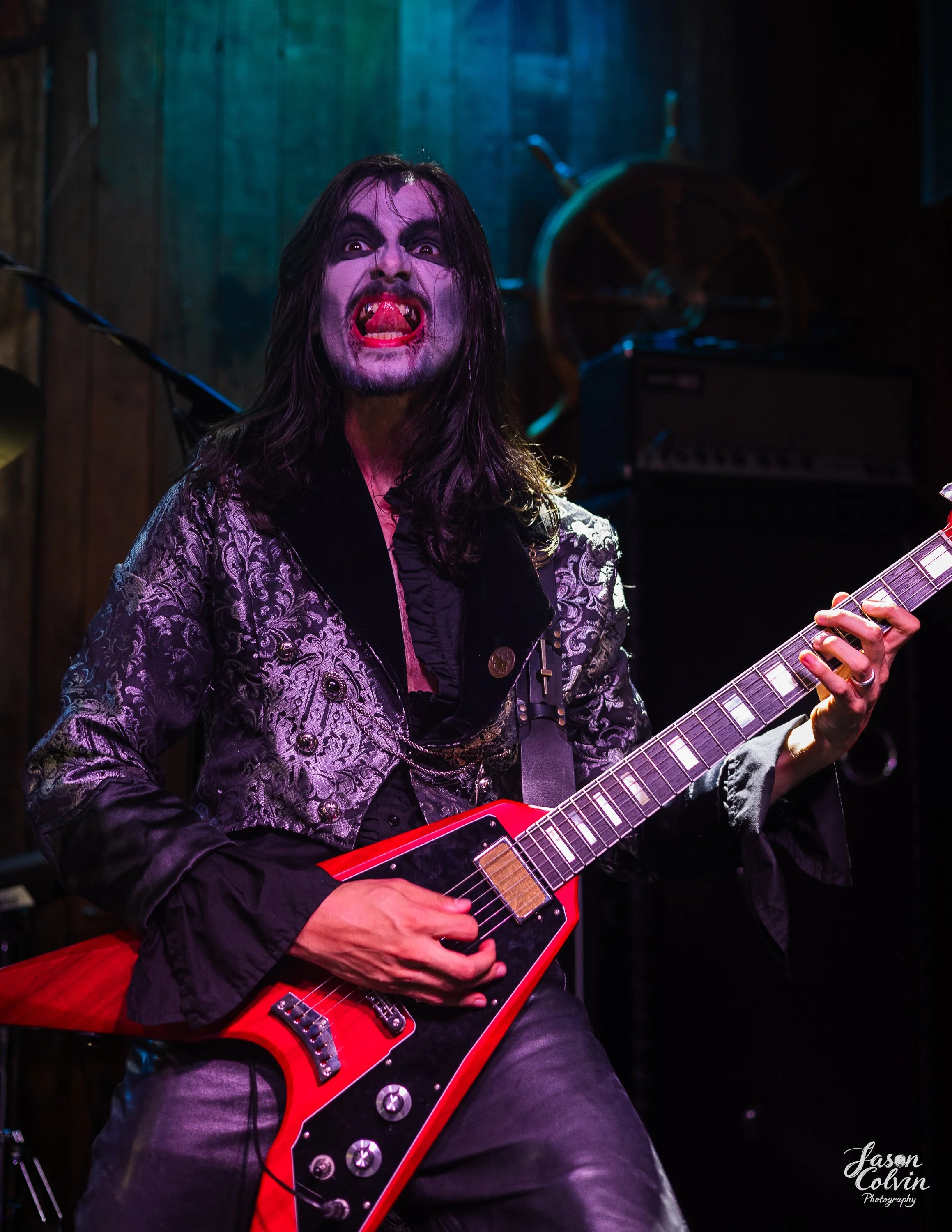A mime dressed as a rock musician is performing live on stage, holding a red electric guitar and making a dramatic face with tongue out, dark makeup, and long black hair.
