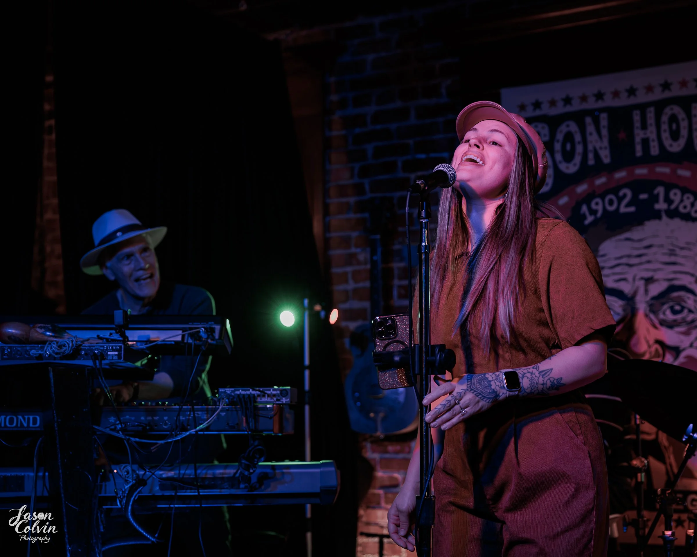 Female singer with long hair and tattoos singing into a microphone, wearing a brown shirt, and a tan hat, with DJ playing music in the background in a dimly lit venue with brick walls.