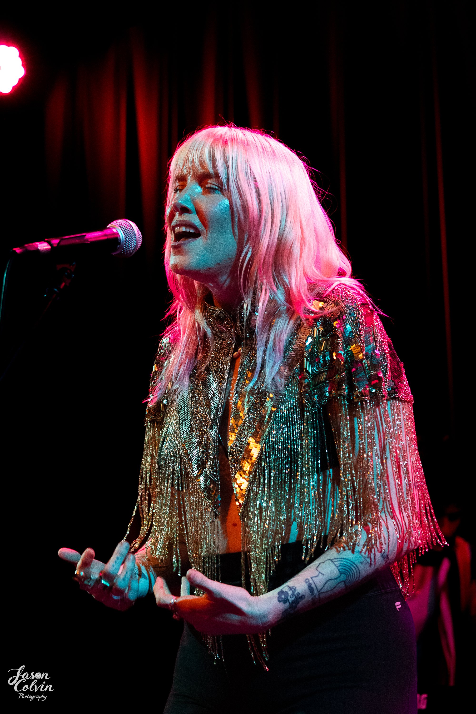 A female singer performing on stage with a pink and blue lighting, wearing a glittery, fringed jacket and singing into a microphone.