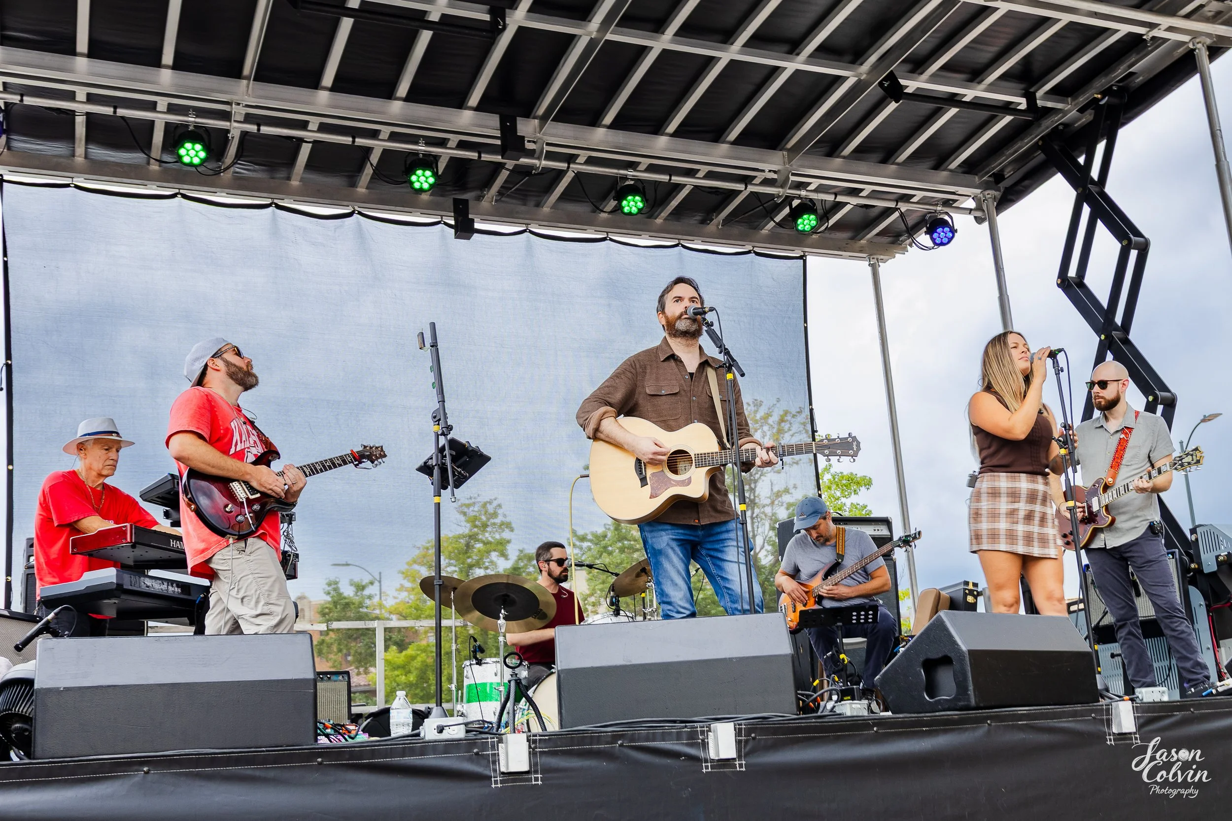 A band performing on an outdoor stage with six members playing various instruments, including guitars, keyboard, and drums, under a black canopy with green and purple stage lights, during daytime with a partly cloudy sky and trees in the background.