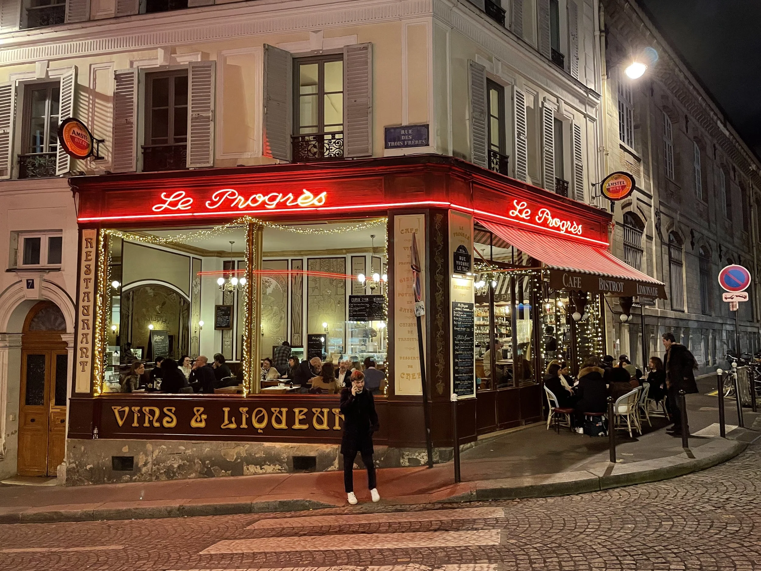 Nighttime view of a corner cafe named Le Progrès with outdoor seating, strings of lights, and a bright red neon sign, with people dining inside and outside on the sidewalk.