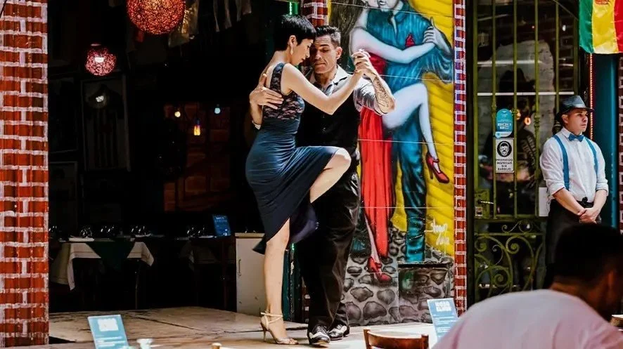 Penguins standing on snow near a calm body of water with icebergs and snow-capped mountains in the background.