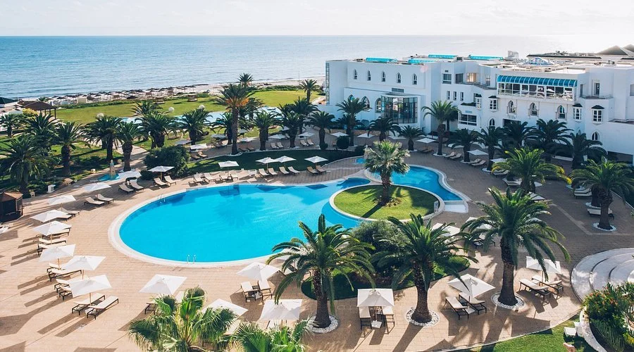 A luxury hotel pool area with lounge chairs and umbrellas, surrounded by palm trees and overlooking the ocean at a sunny day.