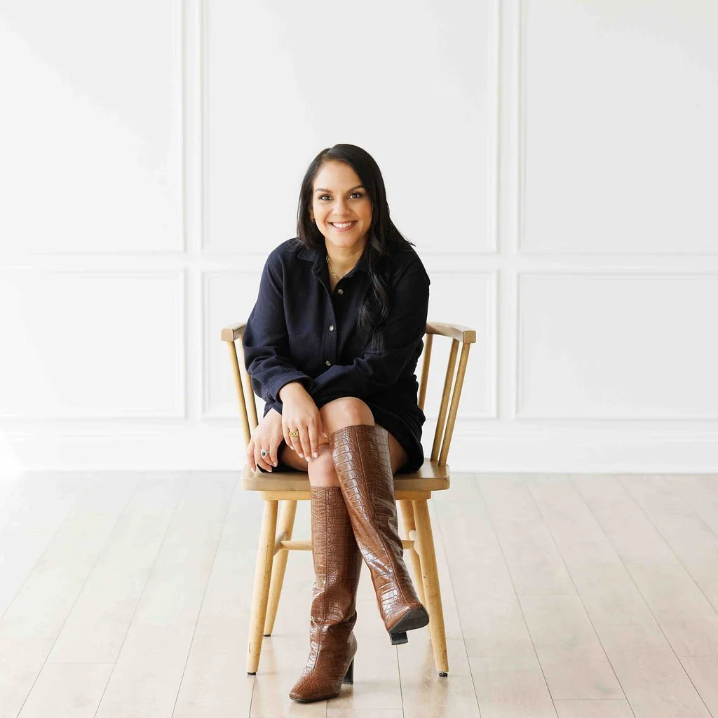 A woman with long dark hair, smiling, sitting on a wooden chair in a bright, white room with paneling, wearing a dark shirt, and brown knee-high boots.
