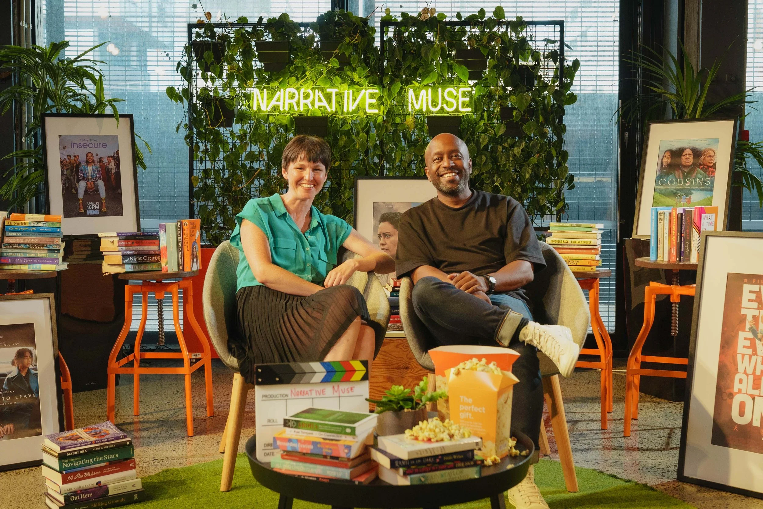 Two people sitting on chairs in a book-filled studio with a neon sign reading 'Narrative Muse' behind them. The woman wears a teal top and black skirt, and the man wears a black shirt and jeans. There are framed posters, stacks of books, and a table with popcorn and a potted plant.
