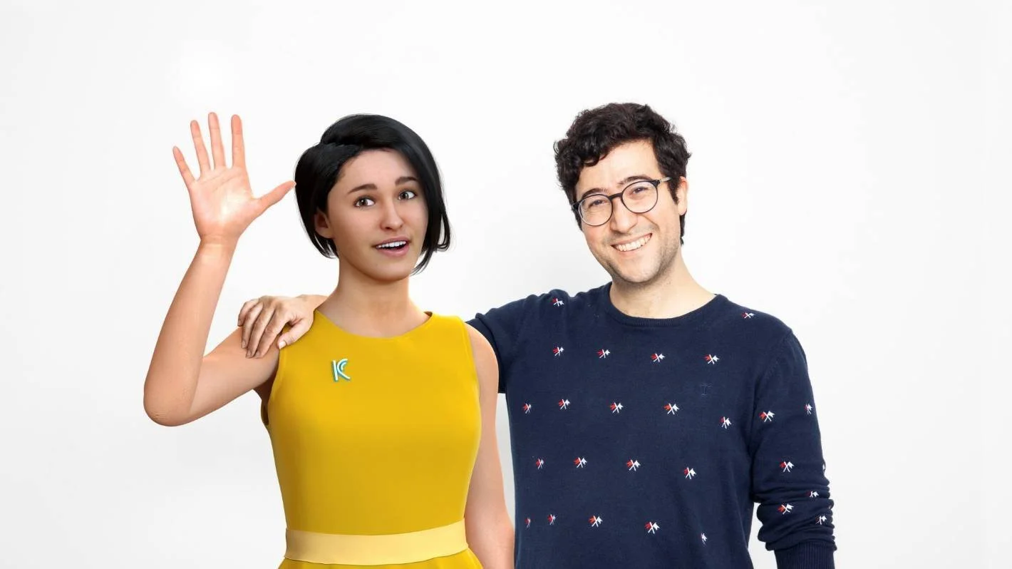 A woman in a yellow dress and a man in a navy blue sweater with small pattern, smiling and waving, standing against a plain white background.