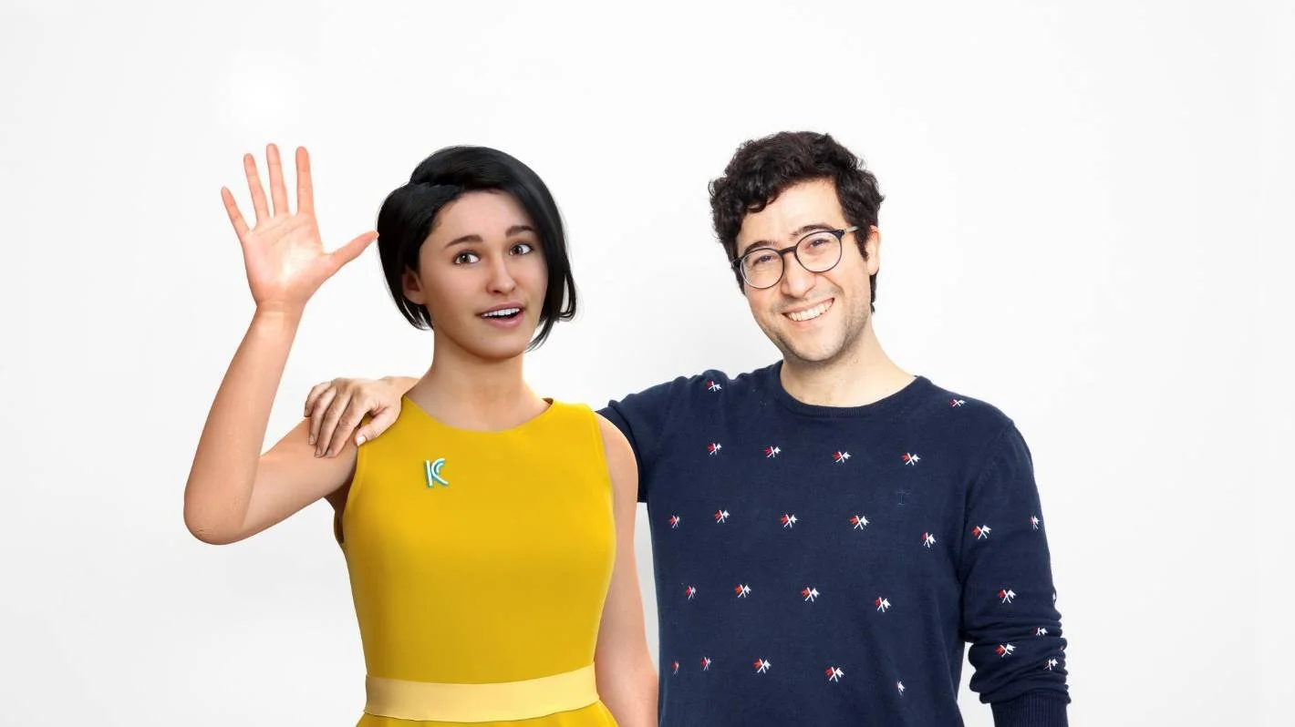 A woman wearing a yellow dress waving and a man in a navy sweater with small flag patterns smiling, standing together against a plain white background.