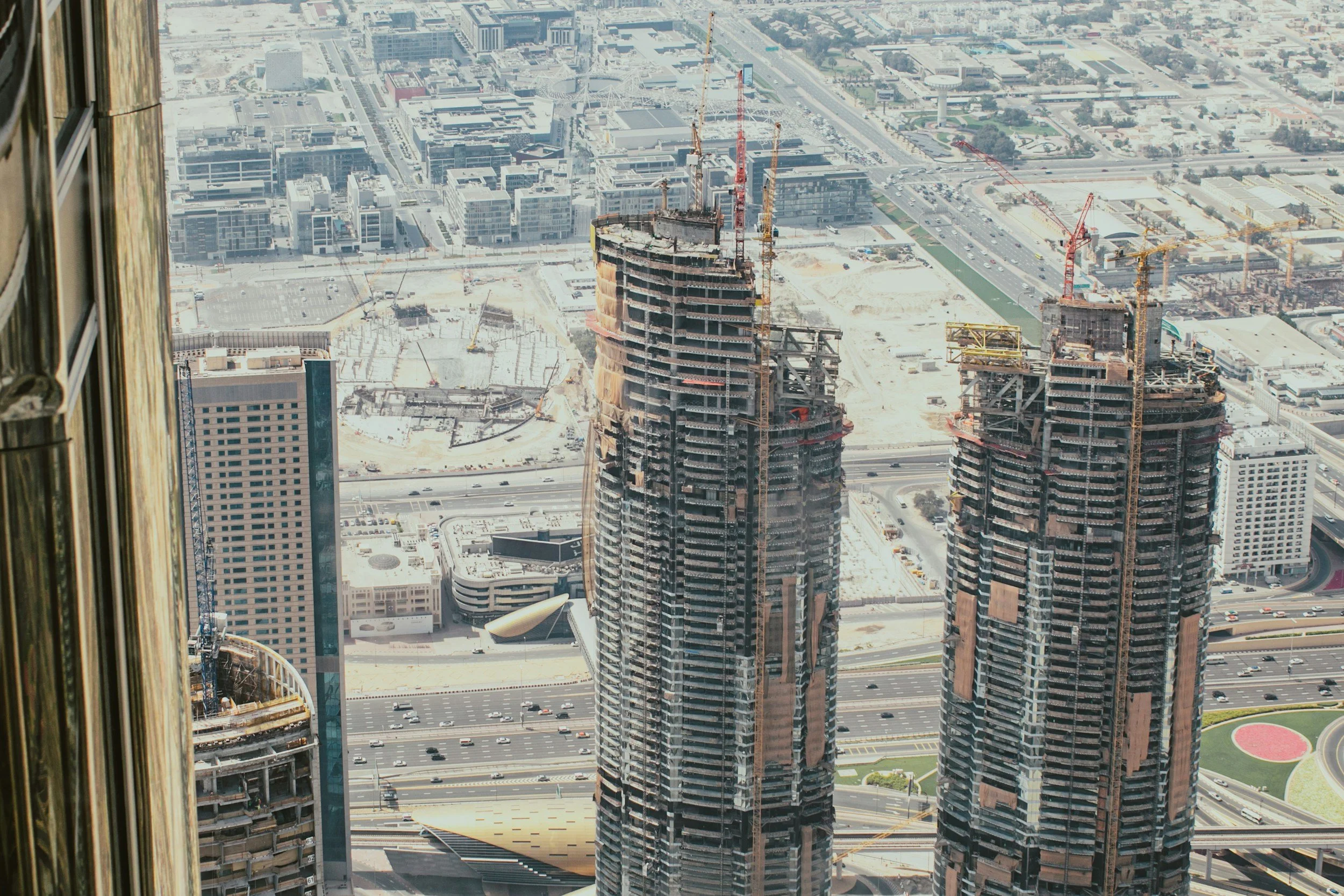 Cityscape view featuring a tall skyscraper under construction with cranes on the building. Other high-rise buildings are visible in the background along with streets, cars, and various city structures.
