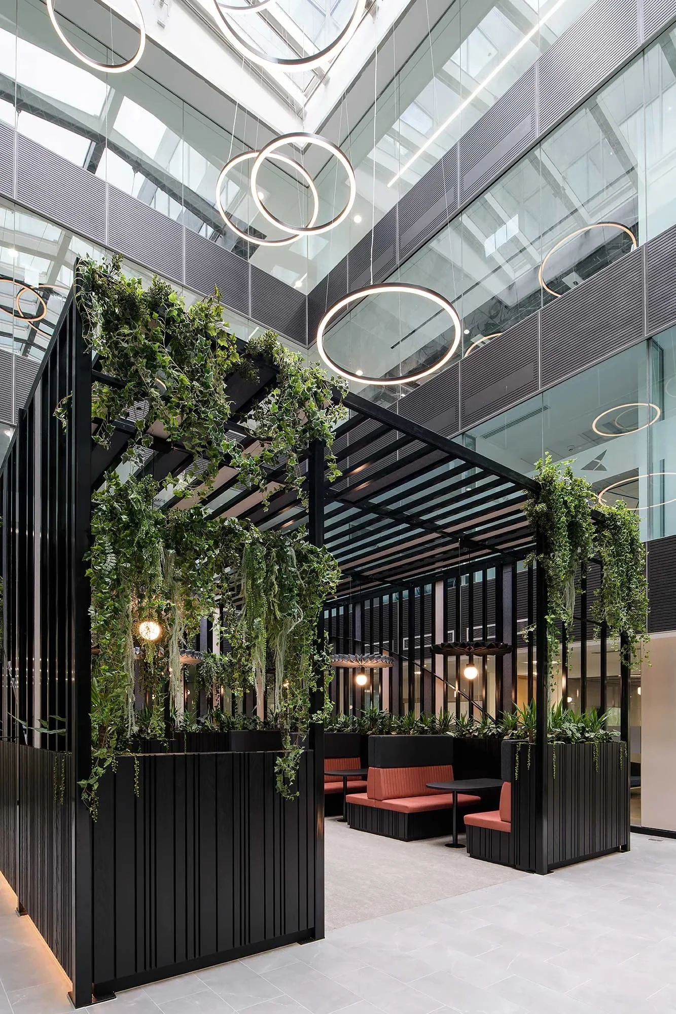 Interior of a modern office or lobby with black metal partition, hanging circular lights, and plant decorations, featuring a seating area with pink chairs and black tables.
