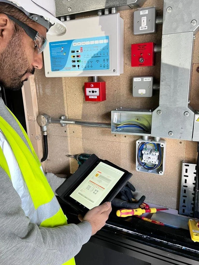 A man in safety glasses and a high-visibility vest working on electrical equipment. He is using a tablet to examine wiring or electrical systems on a wall-mounted control panel with various controls and switches.