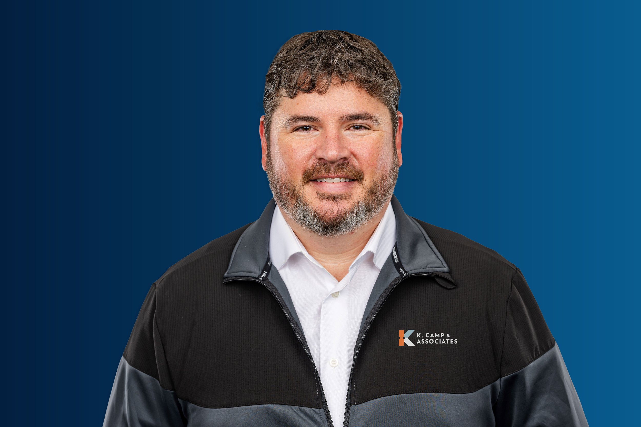 Headshot of a smiling man with curly brown hair and a beard, wearing a black and gray jacket with a logo that reads 'K. CAMP & ASSOCIATES' on the chest, against a blue background.