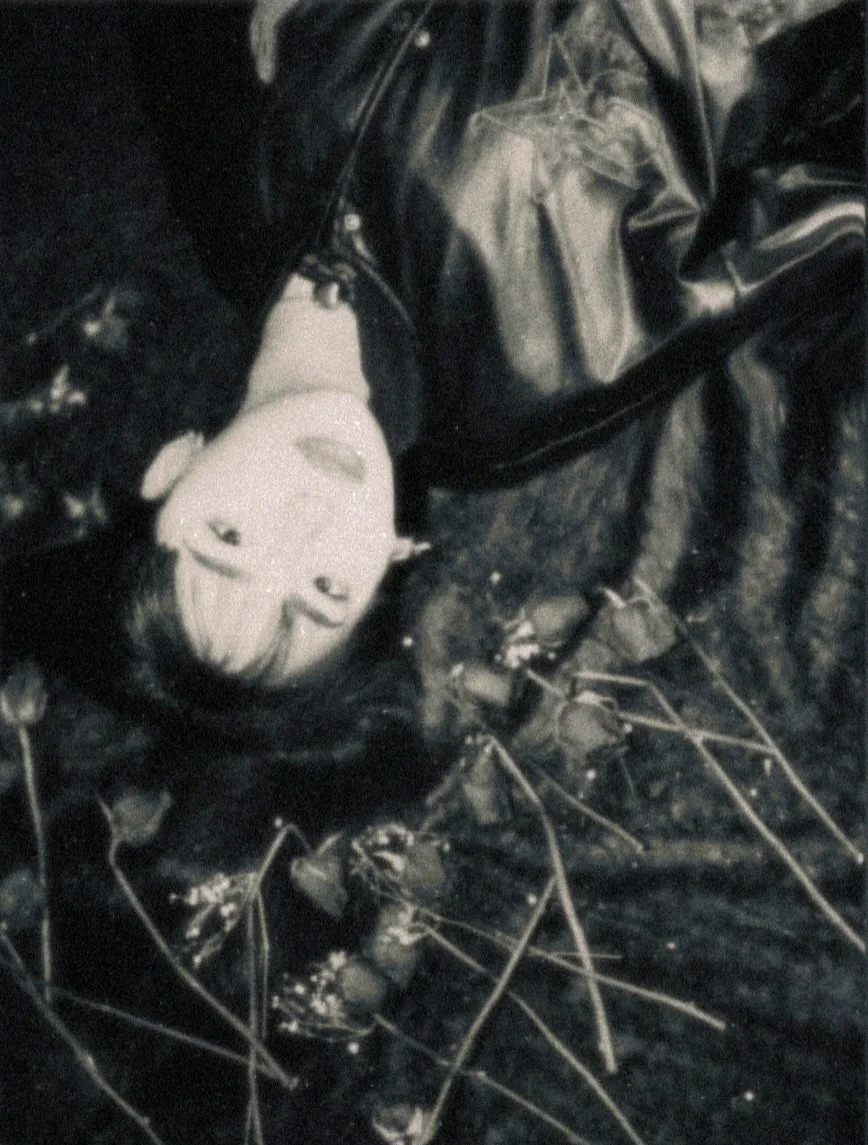 Talia Hilmi lying on the ground in a dark area, surrounded by dried up roses, with a neutral expression on her face.