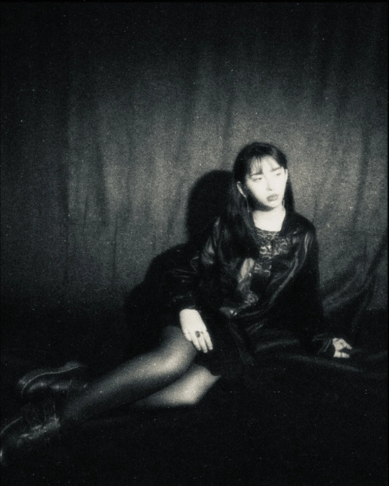 A black and white photograph of Talia Hilmi sitting on the floor against a dark textured wall. She is wearing a leather jacket, lace top, and sheer stockings, with her legs extended and her gaze directed away from the camera.