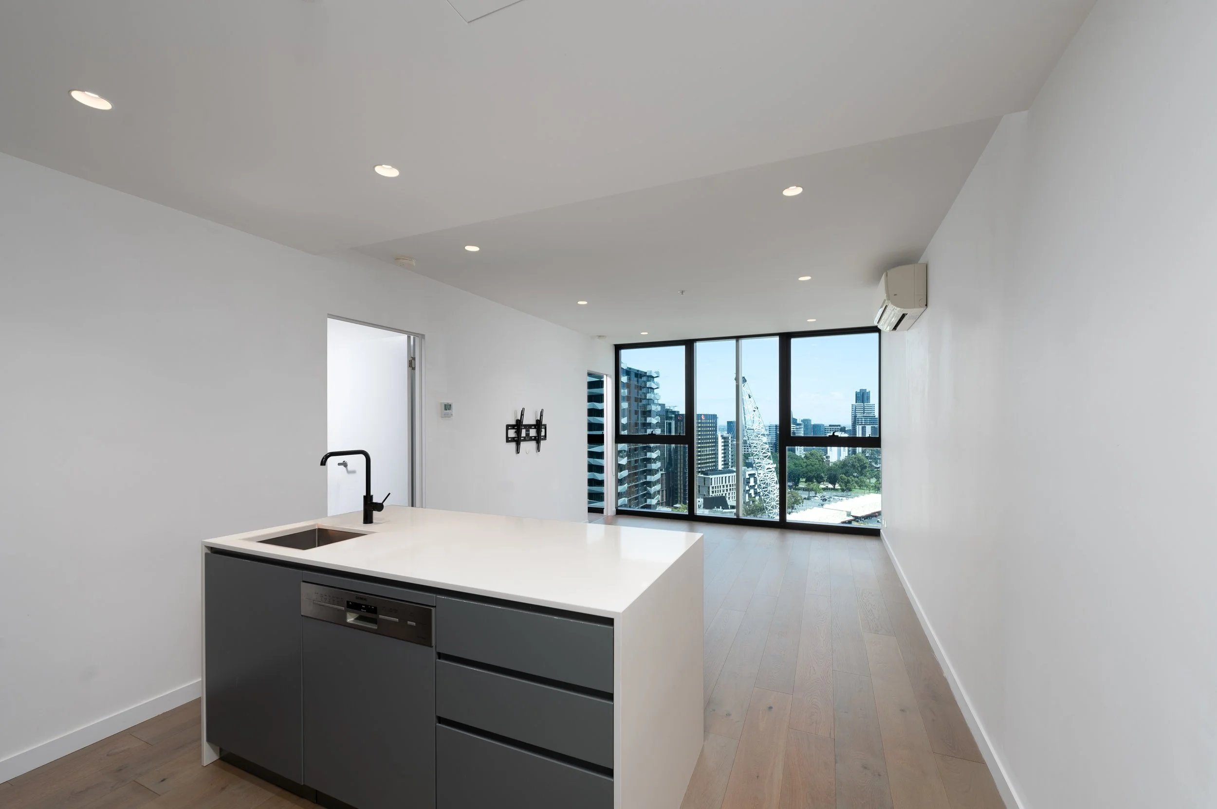 Modern open-plan apartment interior with a kitchen island and sink, large windows offering a cityscape view, and wood flooring.