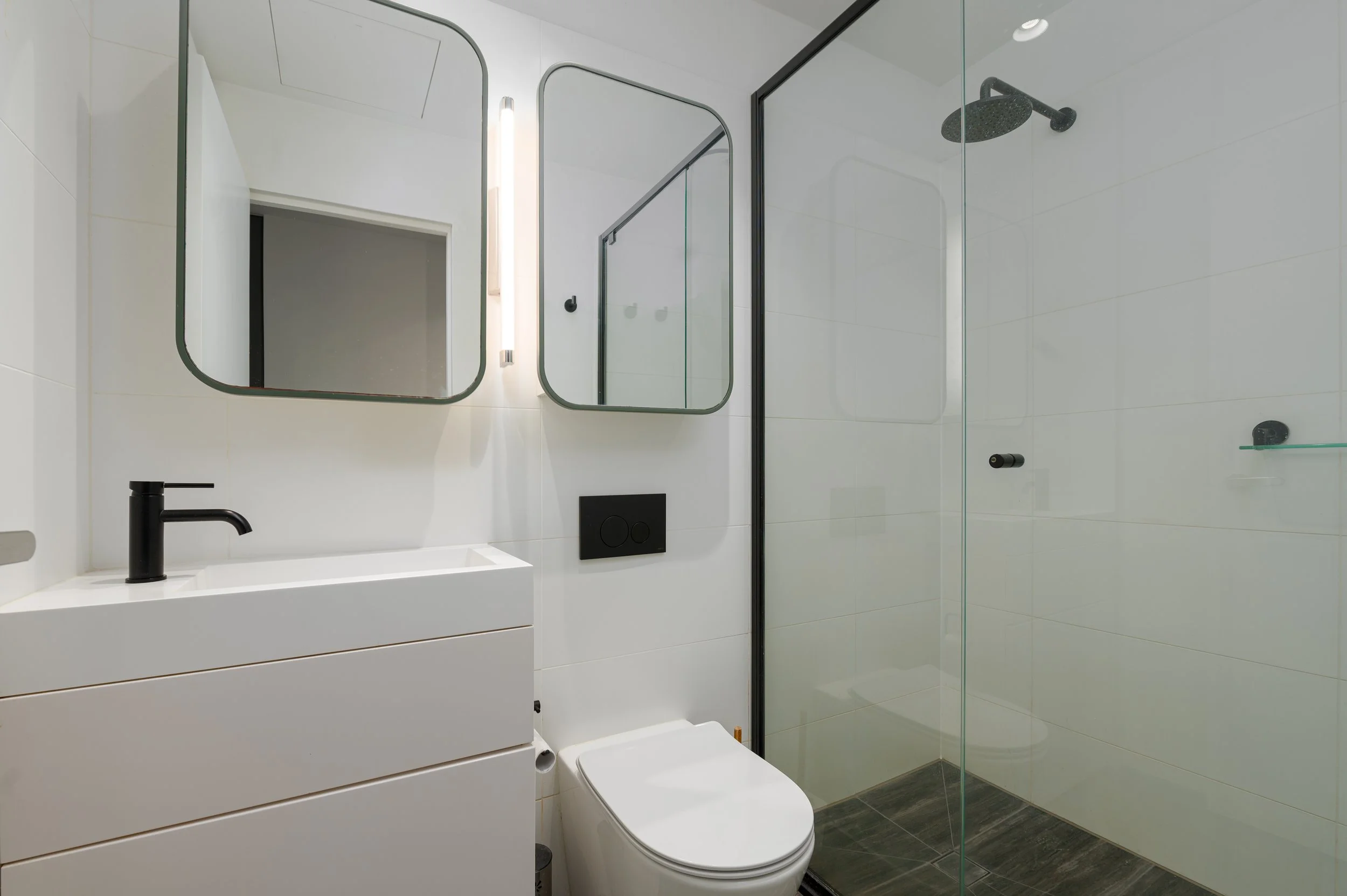 Modern bathroom with white walls, a floating vanity with a black tap, a toilet, glass shower enclosure, and two mirrors.