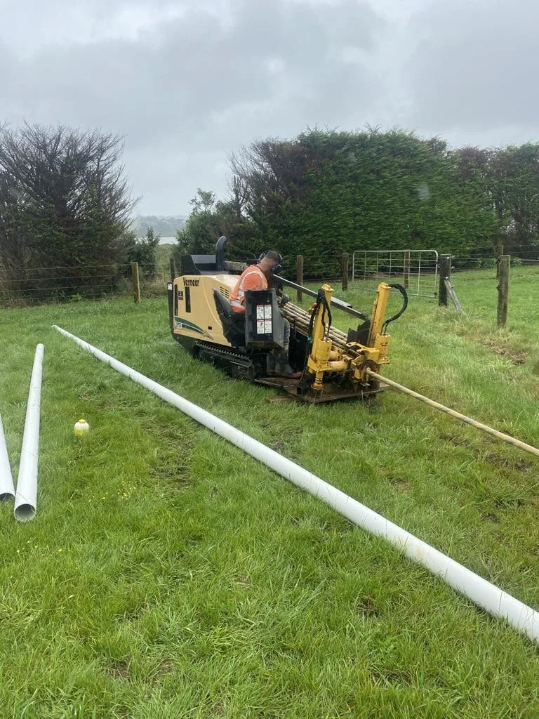 Construction worker using a horizontal directional drilling machine in a grassy field with PVC pipes on the ground.